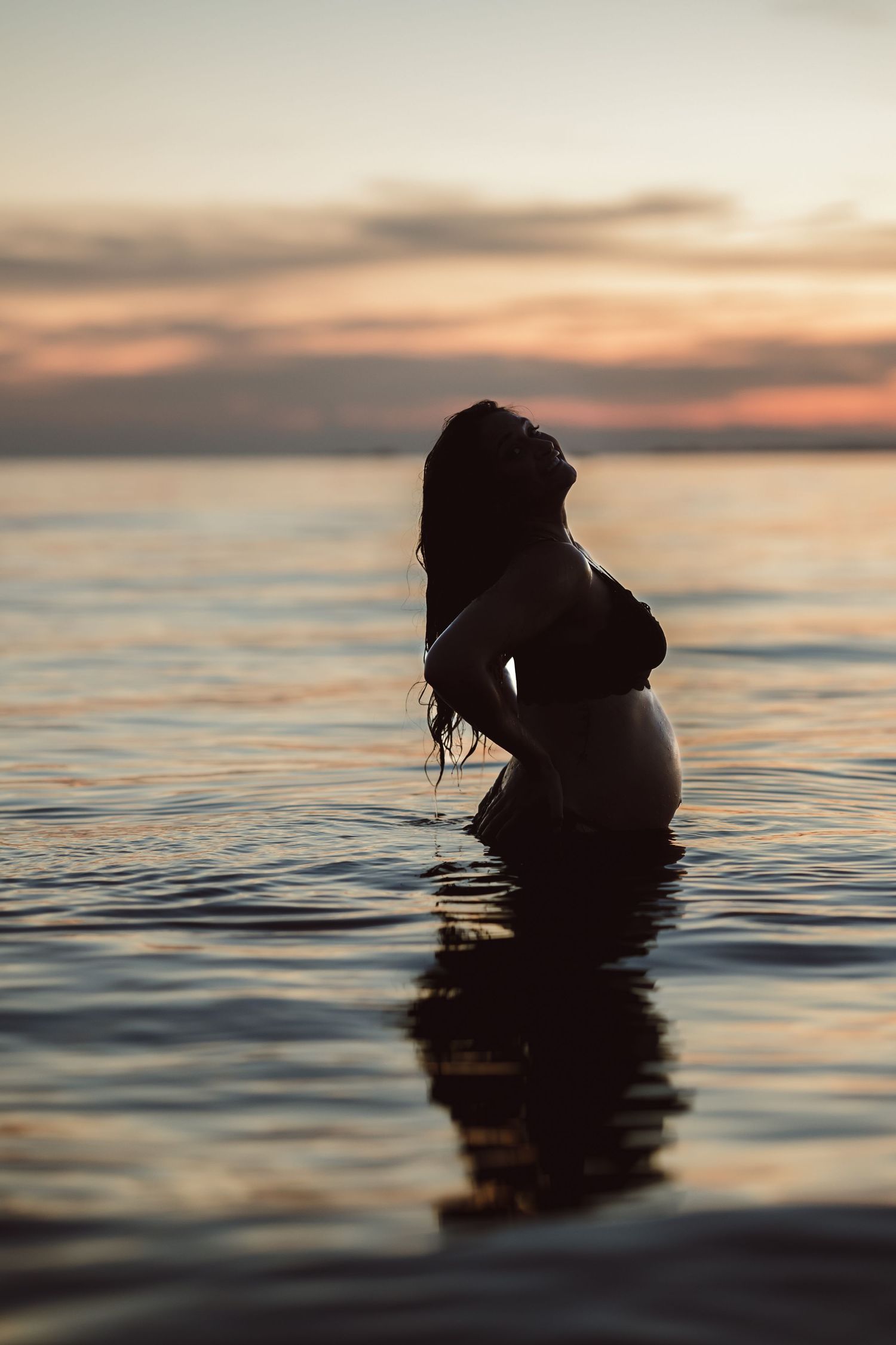Silhouette of pregnant woman standing in water during gold and pink sunset with reflection on calm ocean surface.