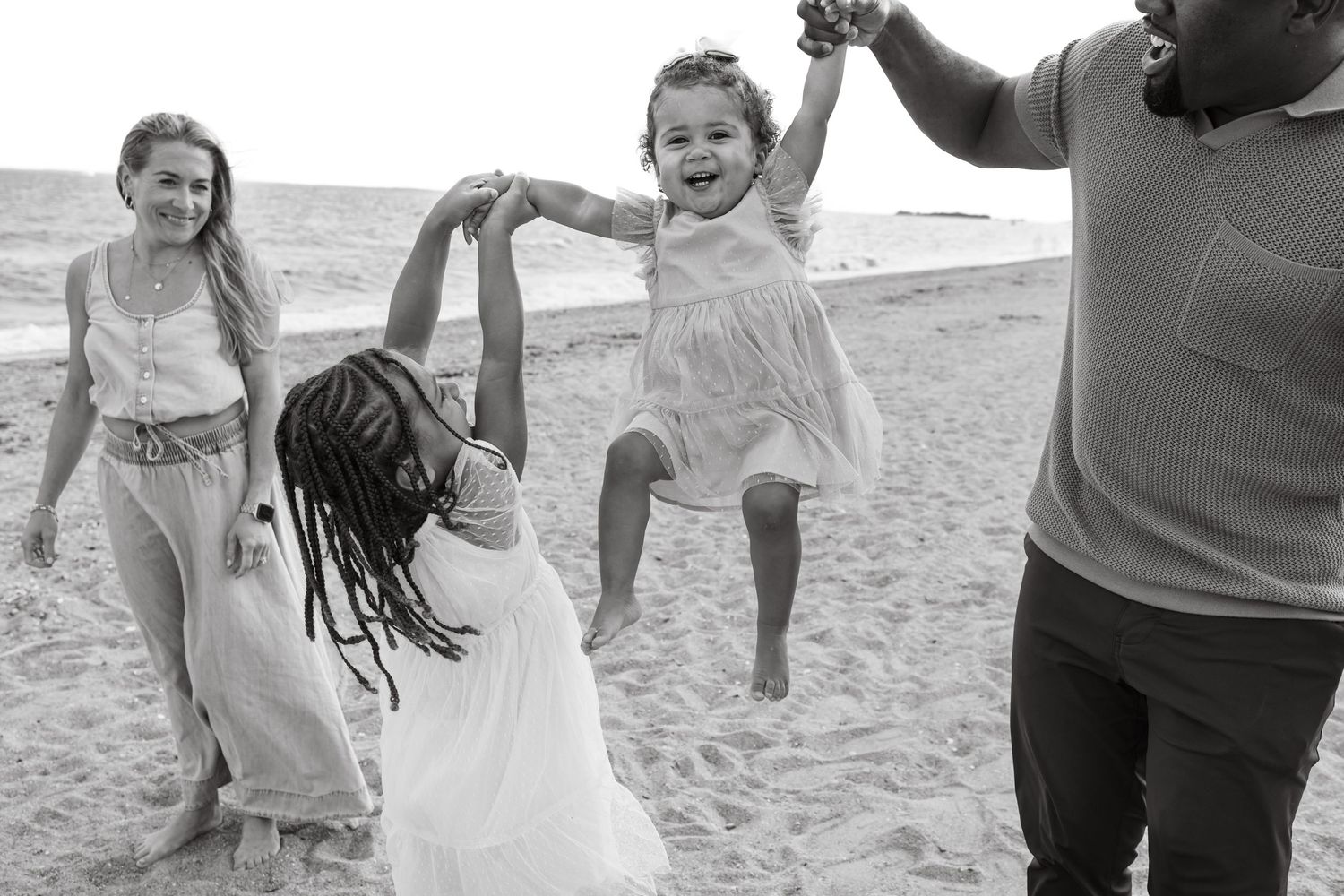 A joyful group plays together on a sandy beach, lifting and dancing in a candid black and white moment.