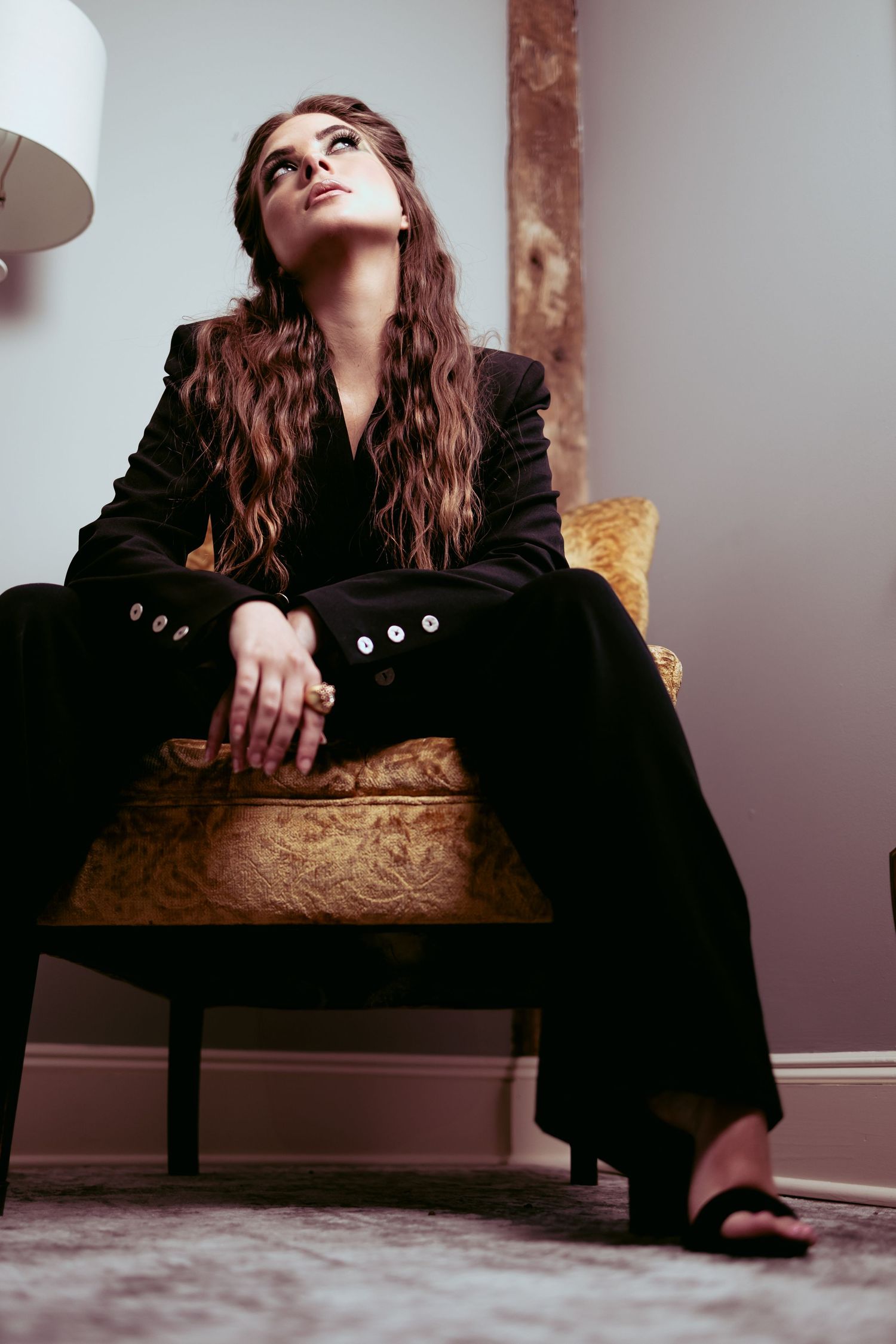 A woman in a black blazer poses dramatically on a vintage chair in a moody studio setting.