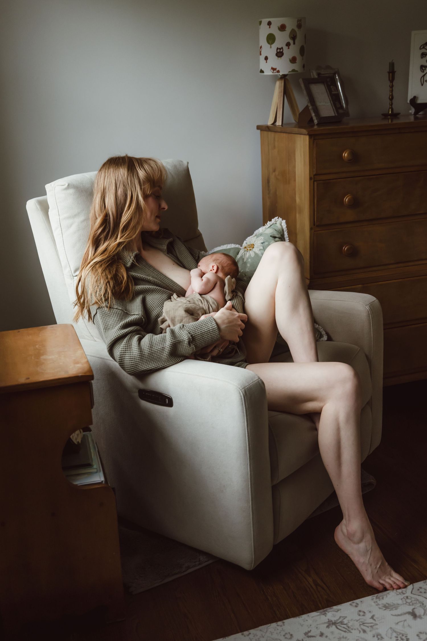 A new mother in a cozy sweater relaxes in a white armchair next to a wooden dresser with a polka dot lamp breastfeeding newborn daughter.