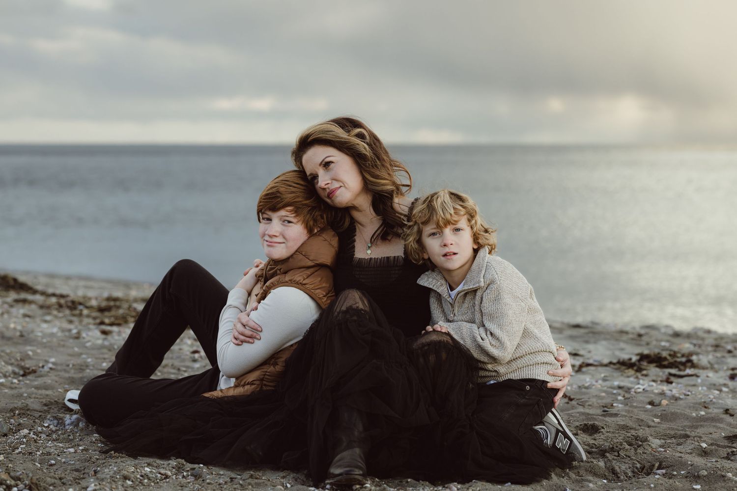 A mother and her two sons embrace each other sitting on the beach at sunset for a motherhood photoshoot.