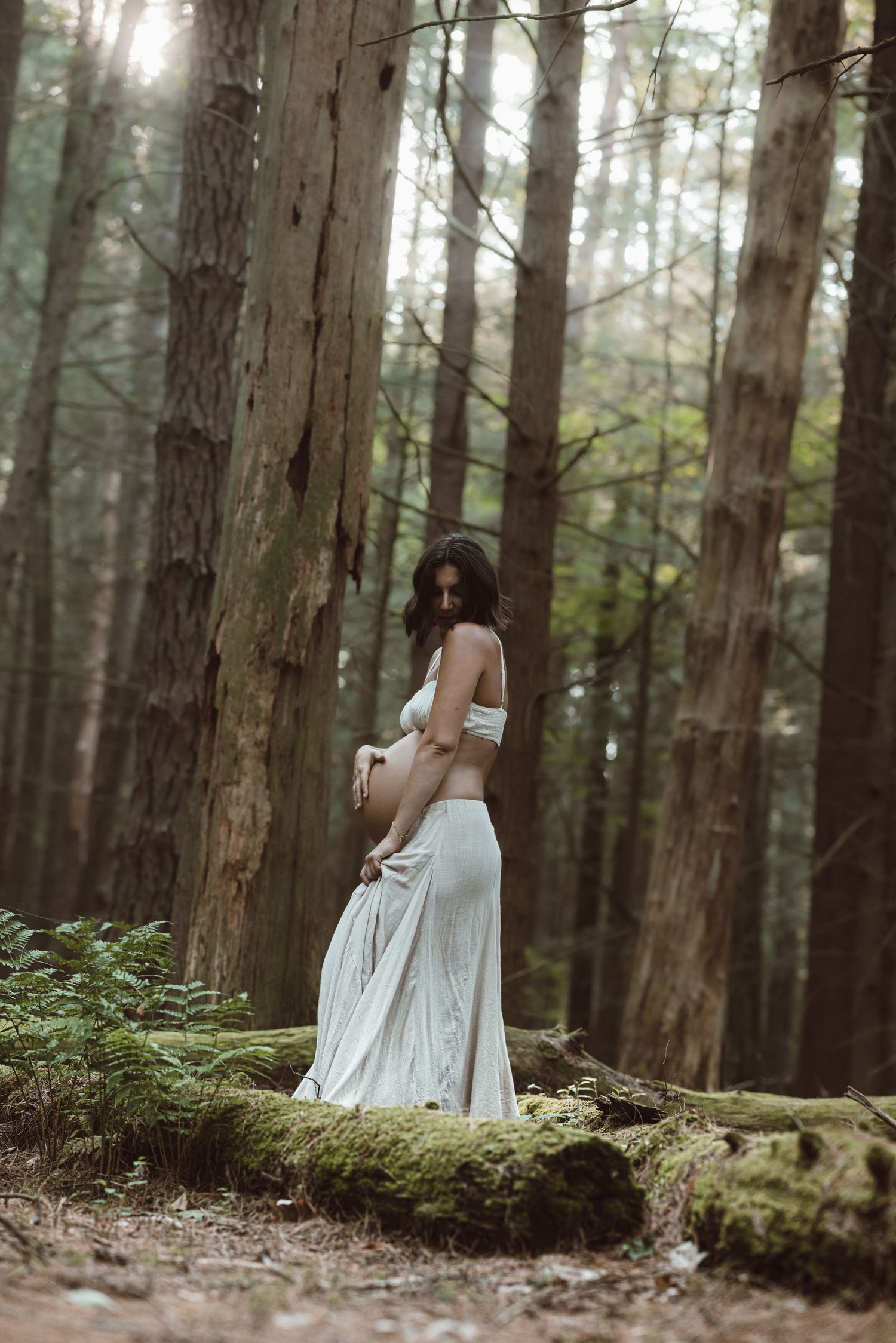 A pregnant woman in a flowing white dress sits ethereally among mossy trees in a misty forest setting for a maternity session.
