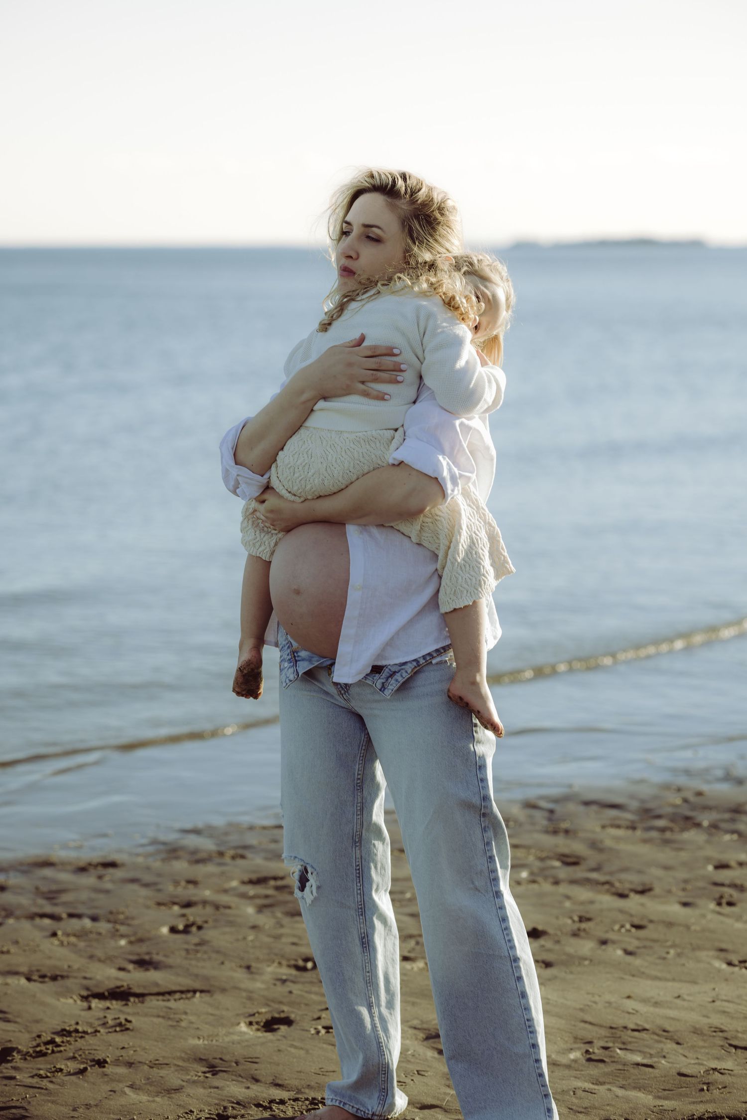 A pregnant mother holds toddler daughter above belly on a Connecticut beach at sunset wearing jeans and an open white shirt.