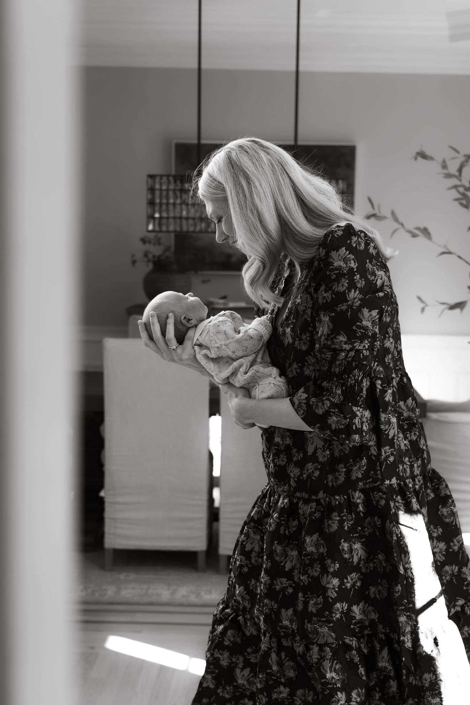 A woman in a floral black dress holds and gazes at a newborn baby in a tender black and white photograph.