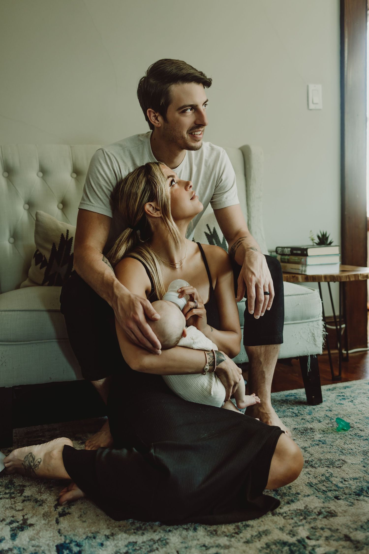 Couple sitting together on floor by tufted couch in living room with patterned rug and potted plant bottle feeding their newborn baby boy.