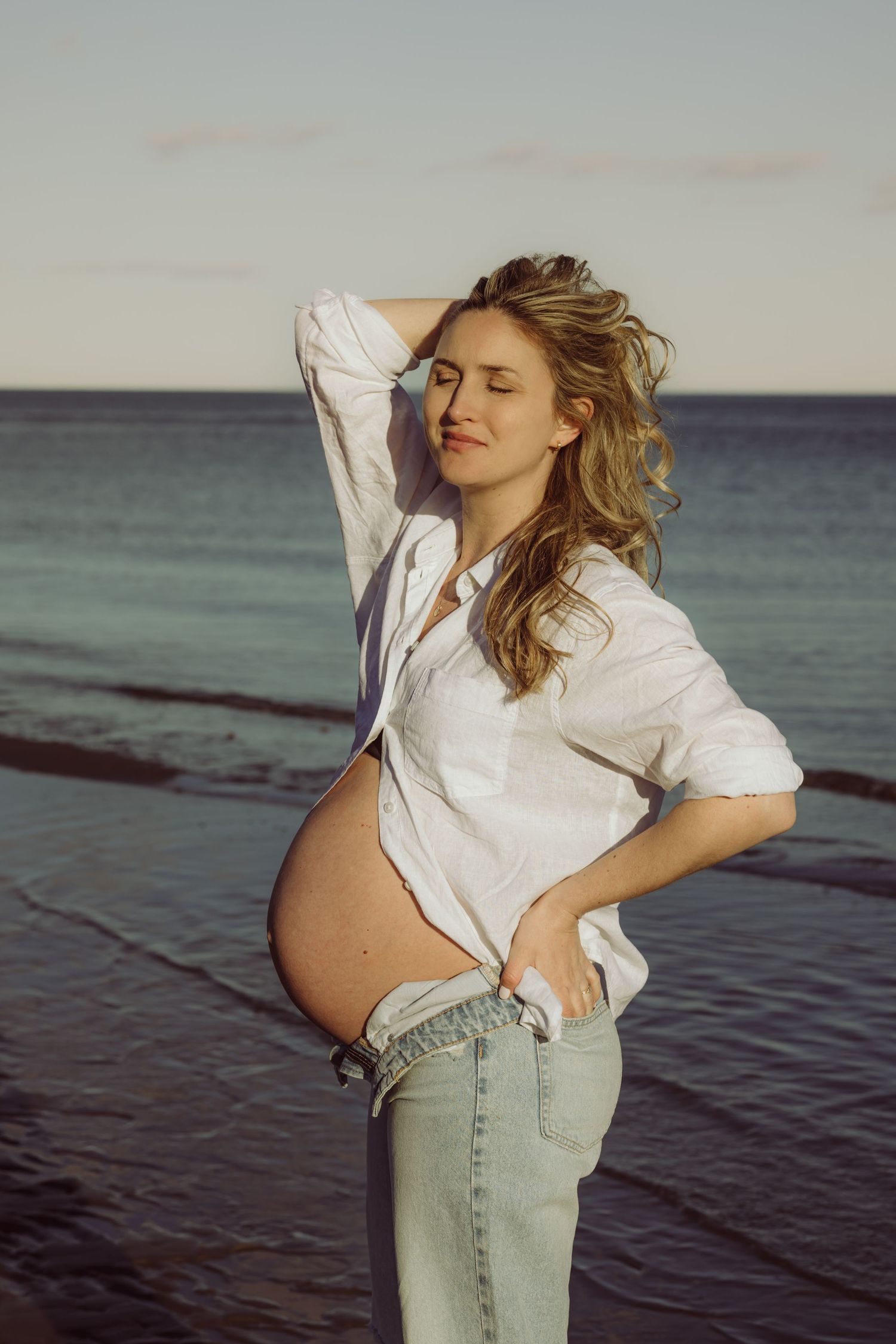 Expectant mother in white button-down shirt and jeans poses on beach at sunset with hair blowing in ocean breeze.