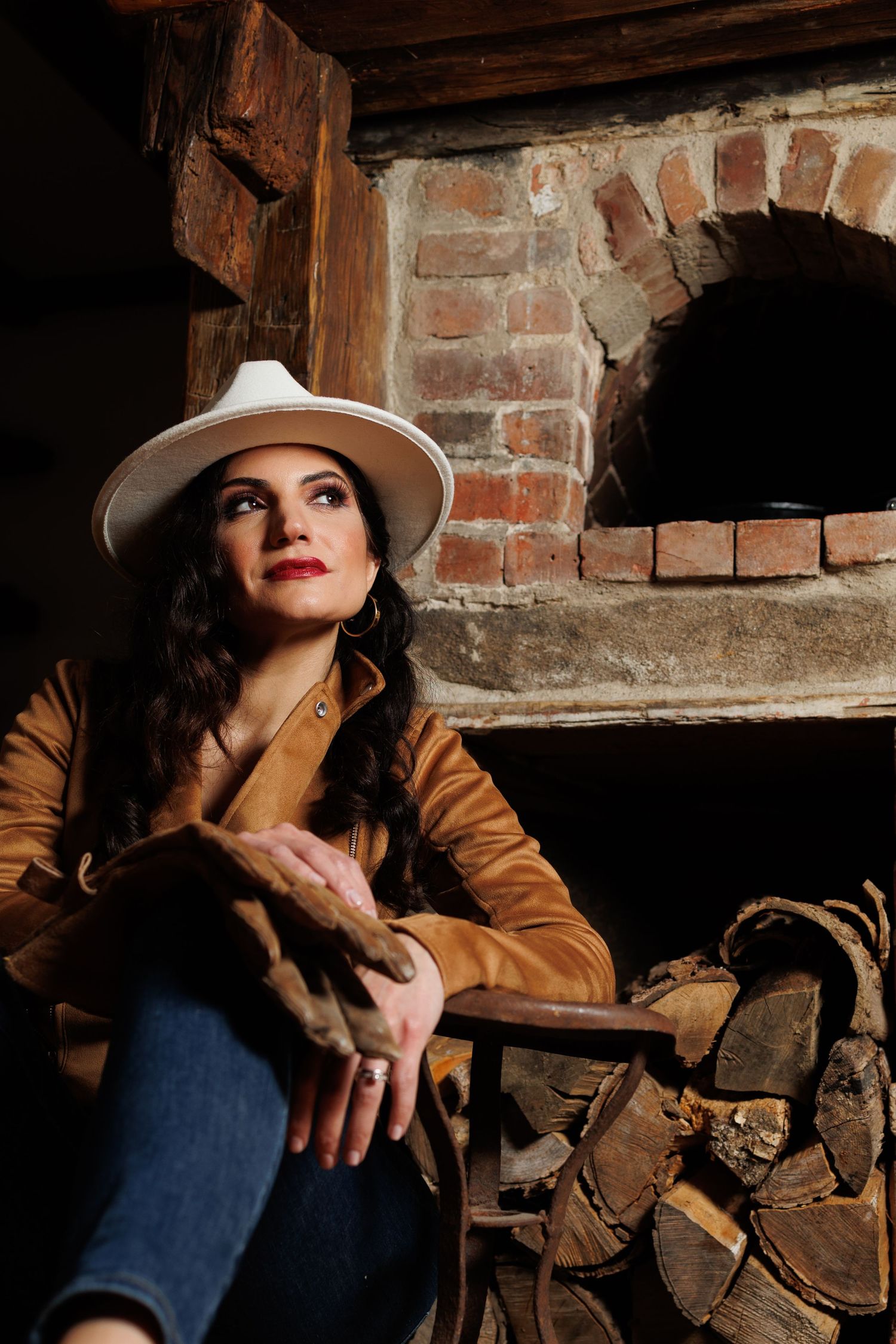 Western-style portrait in rustic setting with white hat, brown leather jacket and denim against brick and wood backdrop.