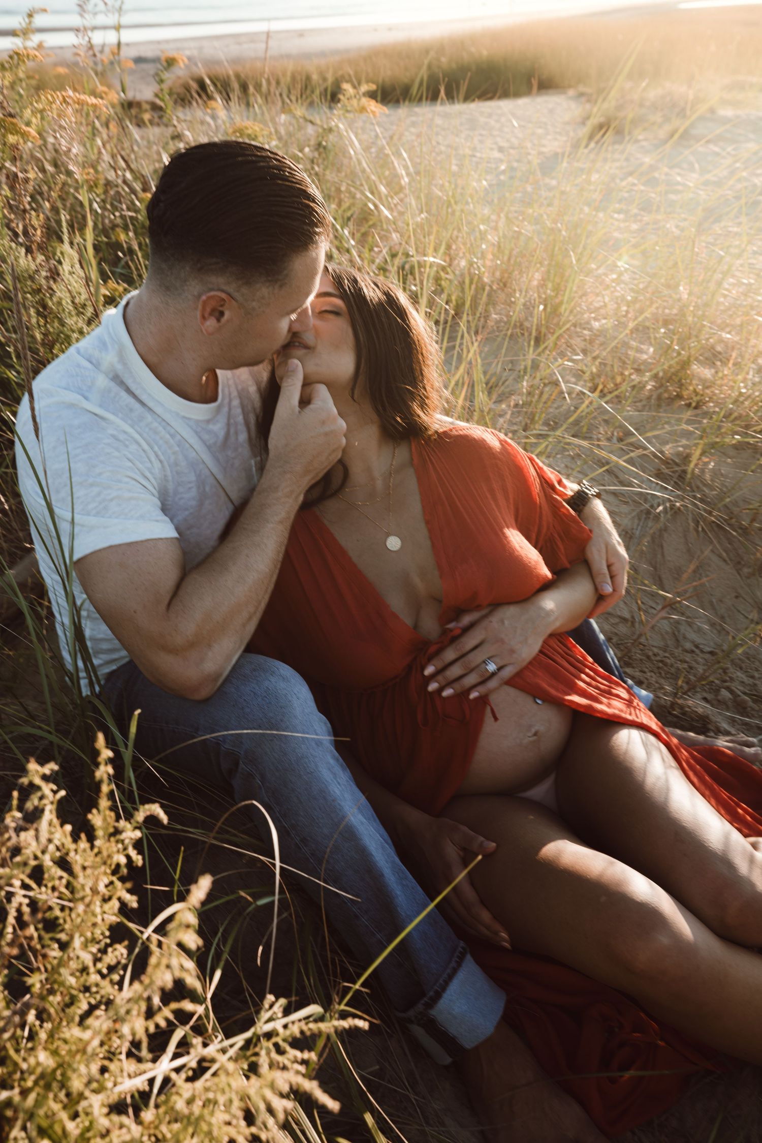 A pregnant woman sitting in between her husbands legs in high grass on the beach at golden hour. He touches her chin and kisses her.