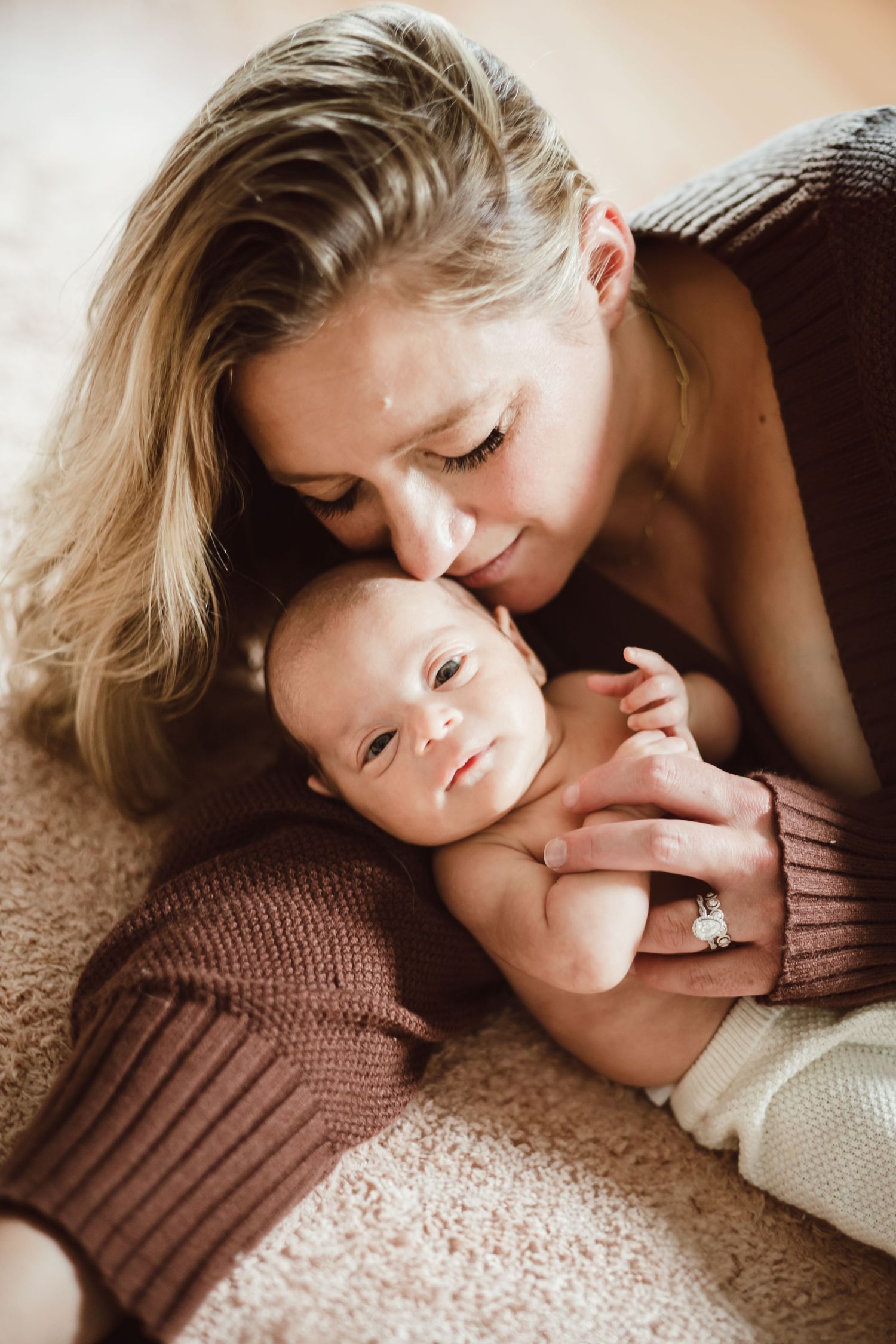 A tender moment captured in warm tones as a mother lovingly kisses their sleeping baby on a textured brown surface.