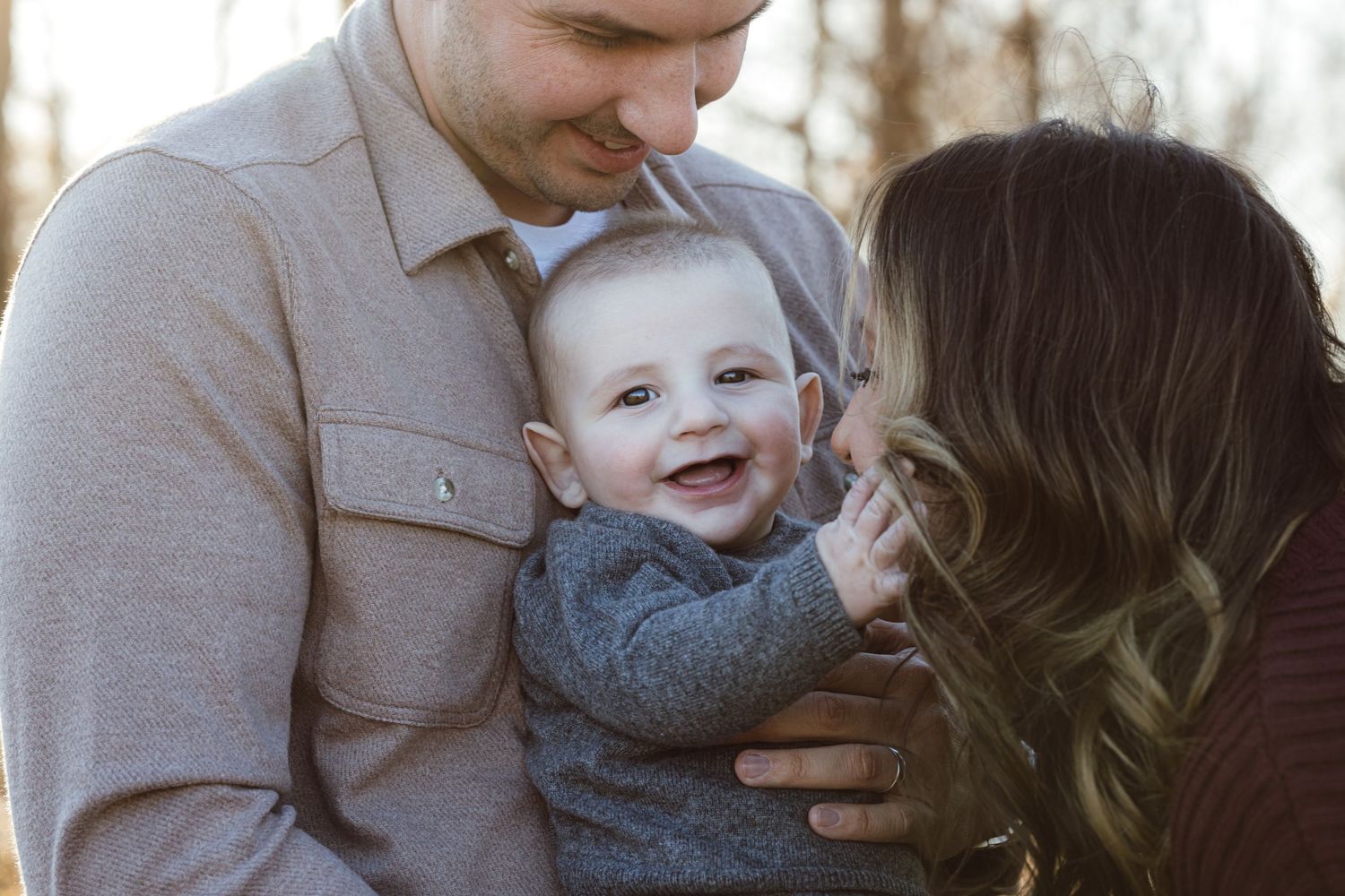 A happy baby in a gray sweater shares a tender moment with parents during an outdoor family photo session.