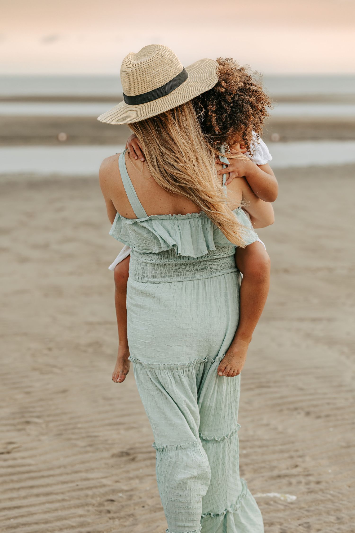 A person in a mint green romper and straw hat walks along a sandy beach at sunset.