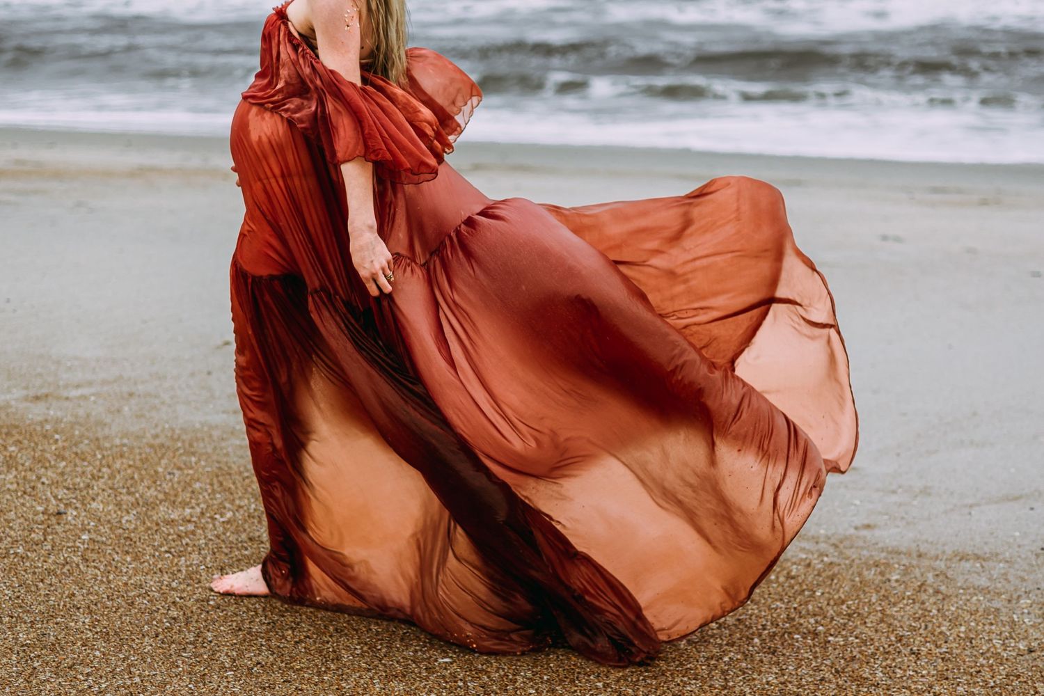 Flowing rust-colored dress billows gracefully in the wind along a misty beach shoreline on a pregnant woman during a maternity photoshoot.
