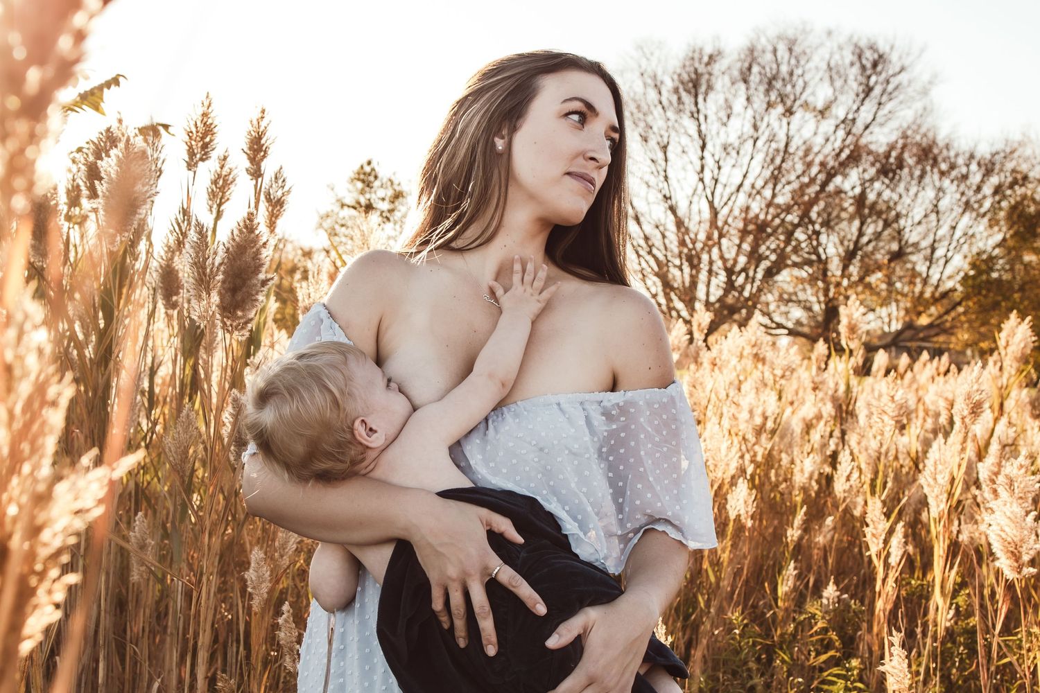 A mother in white dress poses in golden sunlit field with tall grass and trees at sunset breastfeeding her 1 year old.