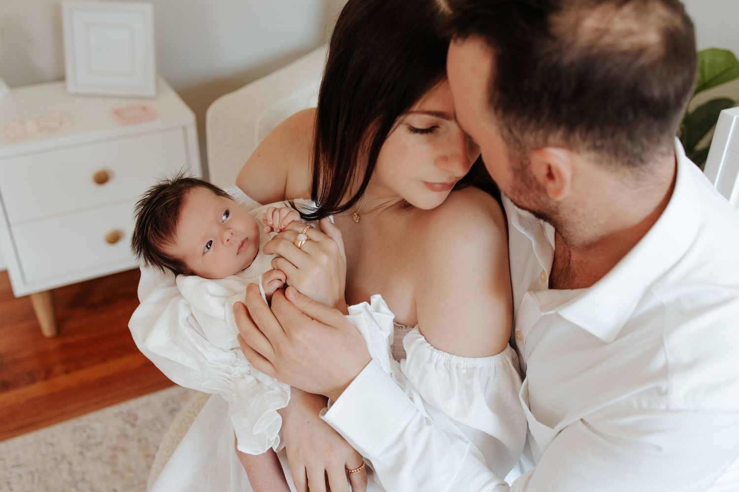 Intimate family moment with newborn baby in white nursery setting with wooden floors and white furniture.