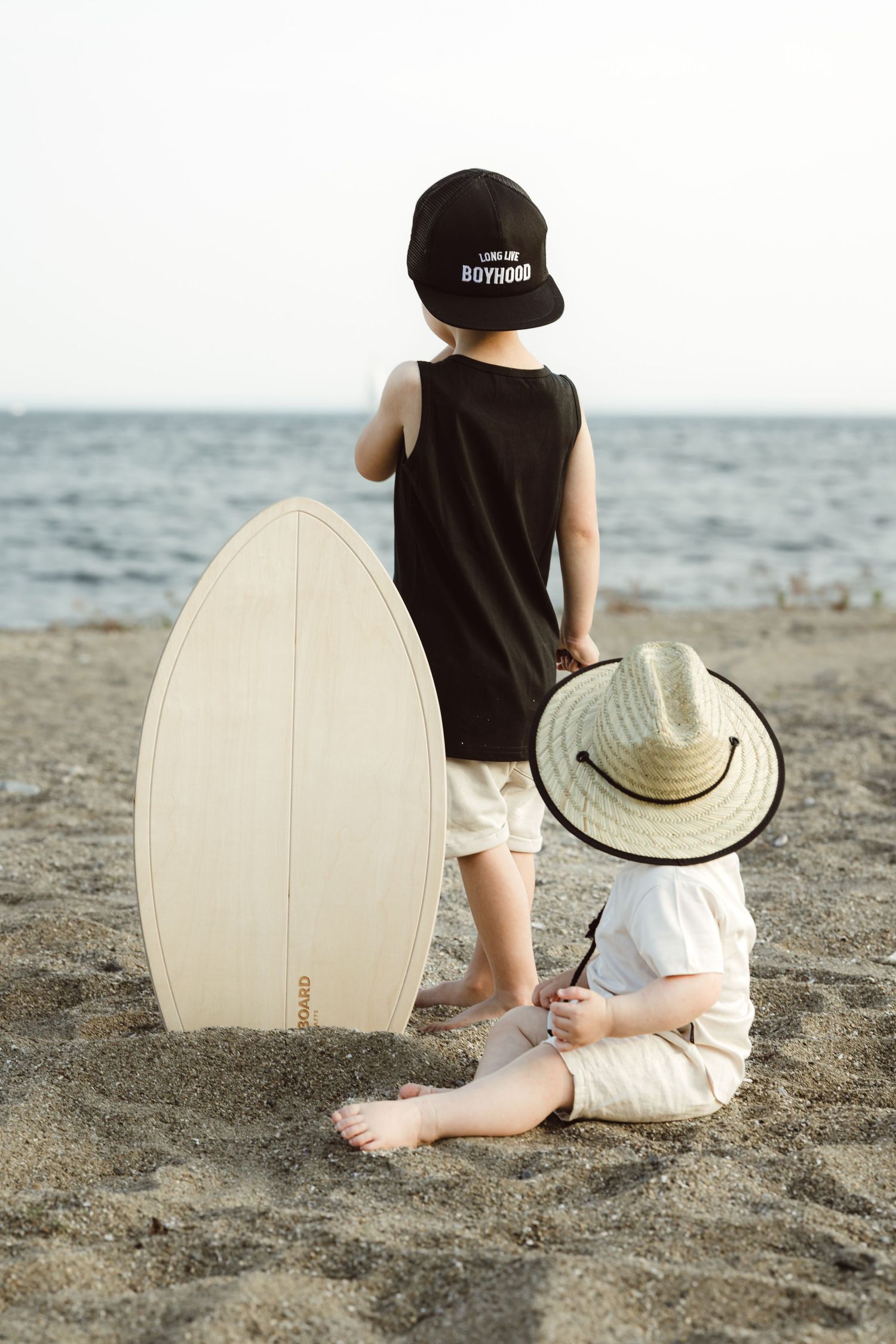 Two boys sit on a sandy beach with a surfboard and straw hat, looking out at the ocean.