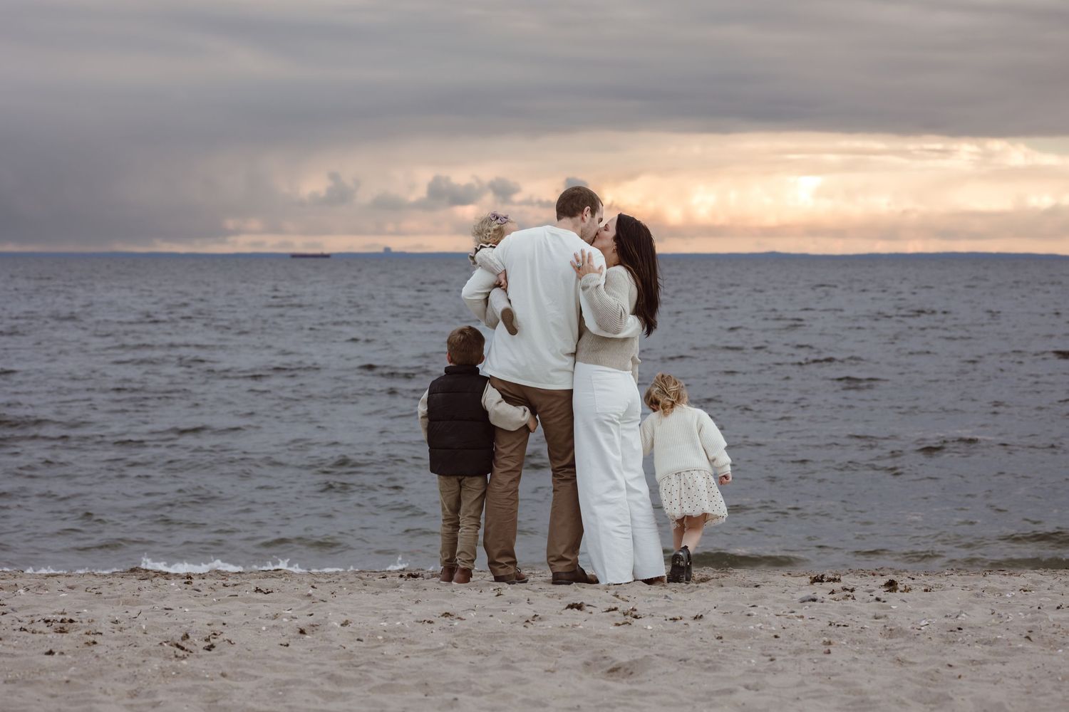 A family stands together on a beach at sunset, silhouetted against the ocean and dramatic cloudy sky.
