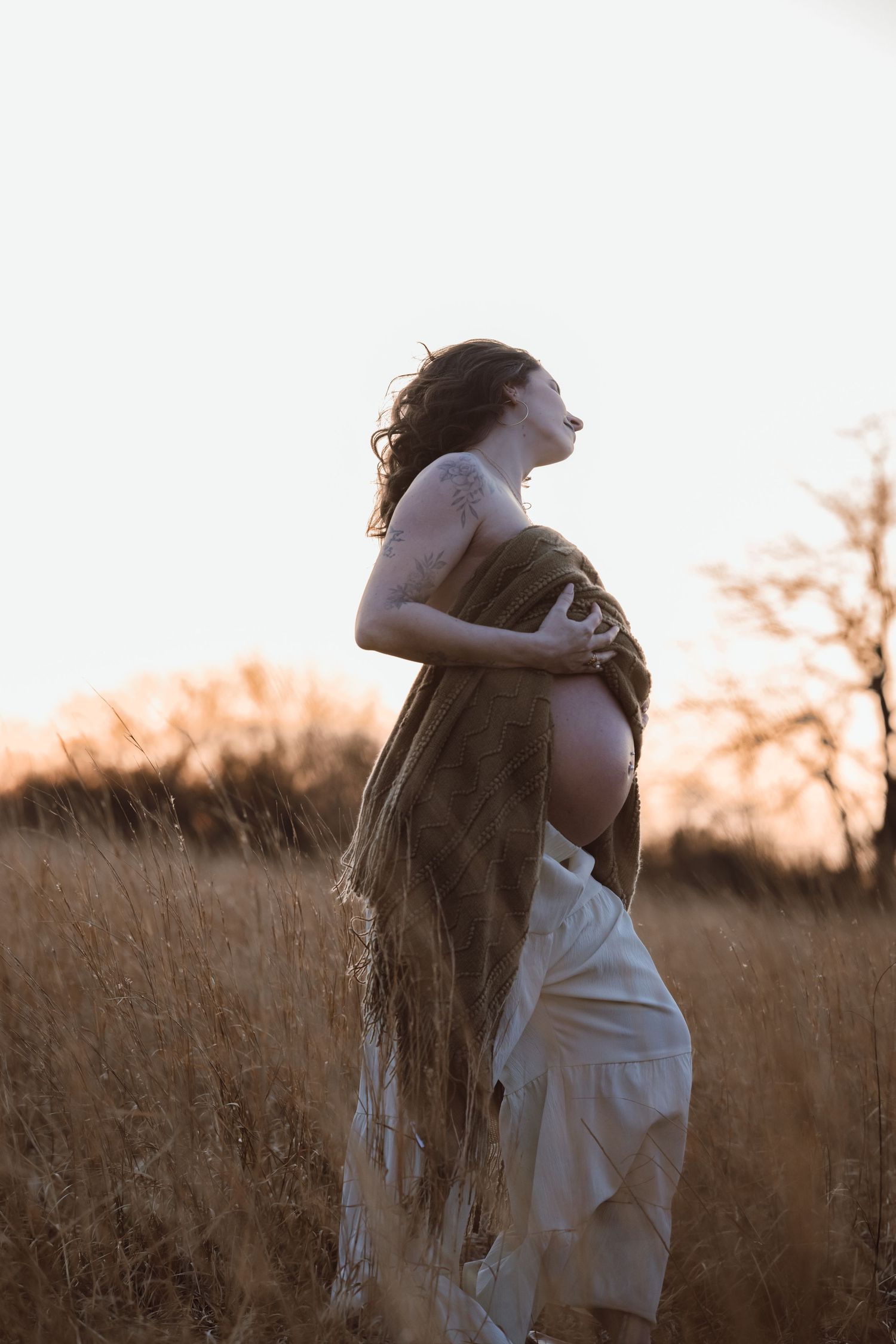 Silhouetted pregnant woman poses with a blanket across chest in a golden wheat field at sunset creating a romantic maternity photo.