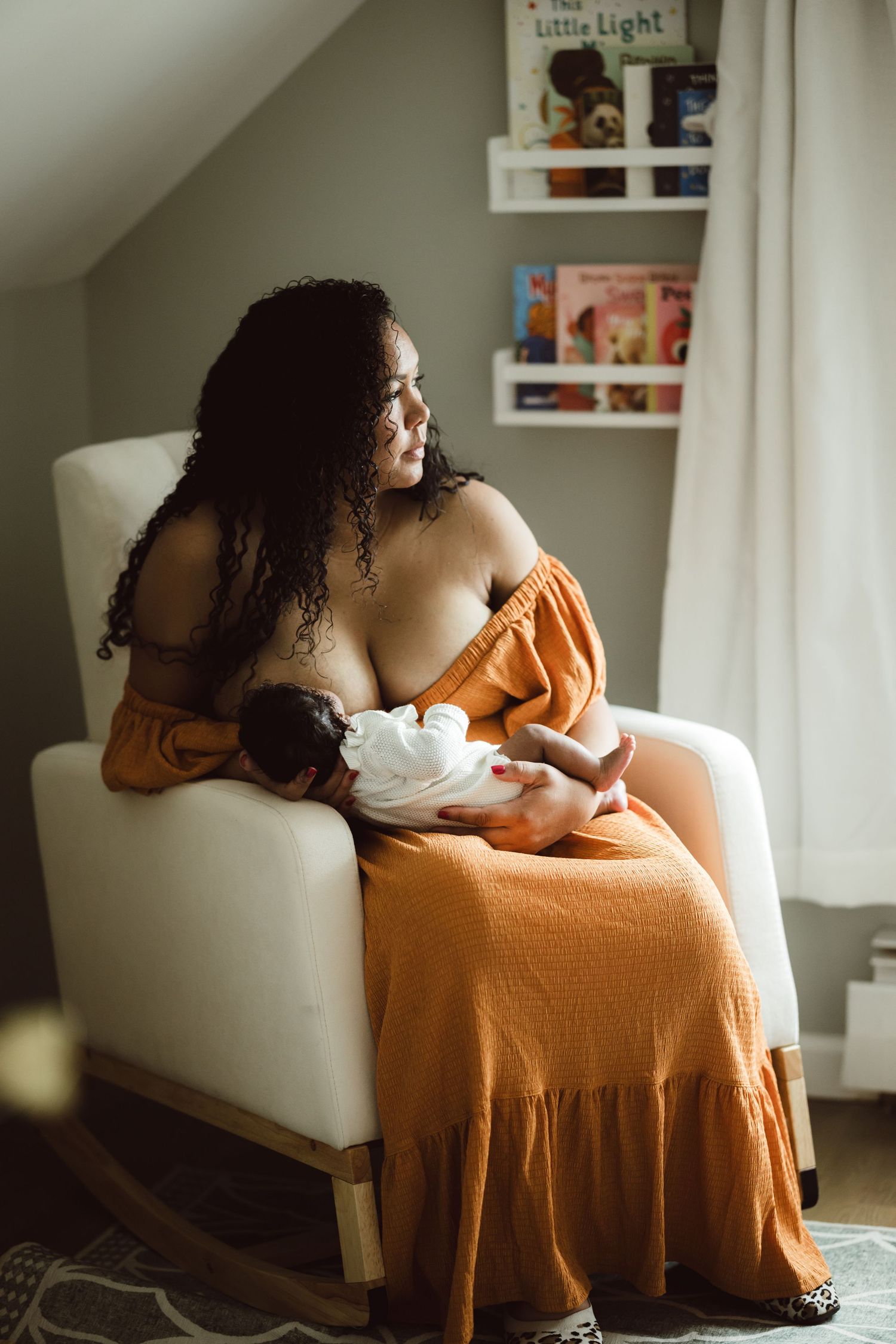 A mom in flowing orange dress lounges gracefully on white chair in sunlit room with bookshelves as she breastfeeds her newborn baby girl.
