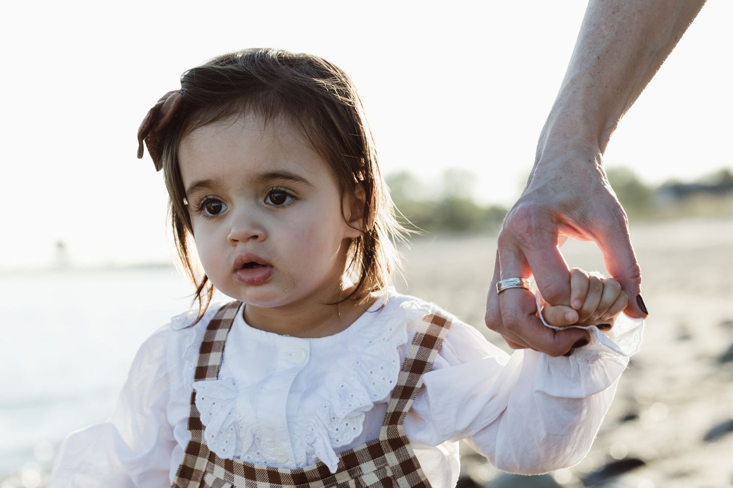 A small child in a white dress holds hands with mother while walking on a sandy beach at sunset for milestone photoshoot.