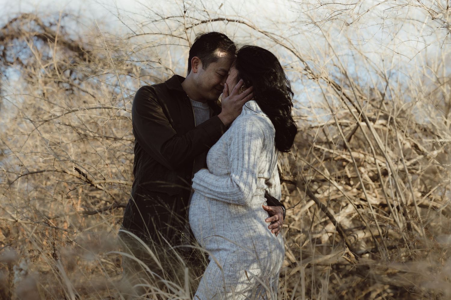 Romantic pregnant couple embraces in a rustic outdoor setting with sunlight filtering through dried branches and foliage for maternity shoot