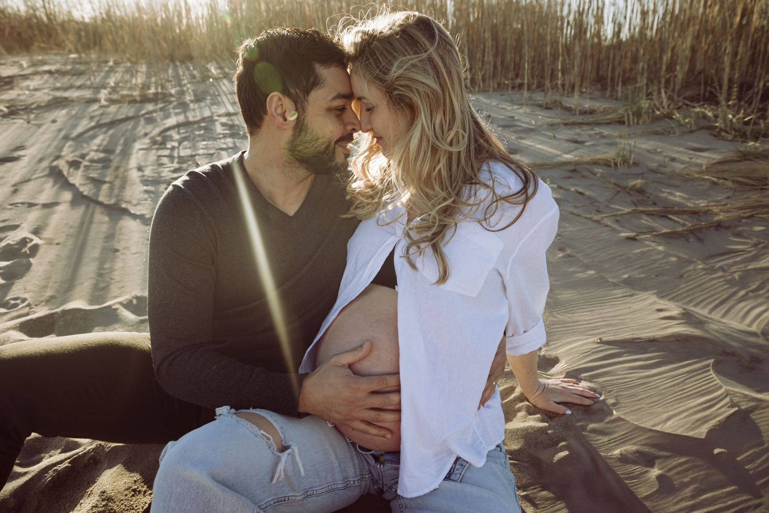 A tender maternity photo on the beach with soft sunlight illuminating a couple sitting in the sand together. Husbands hand on her belly.