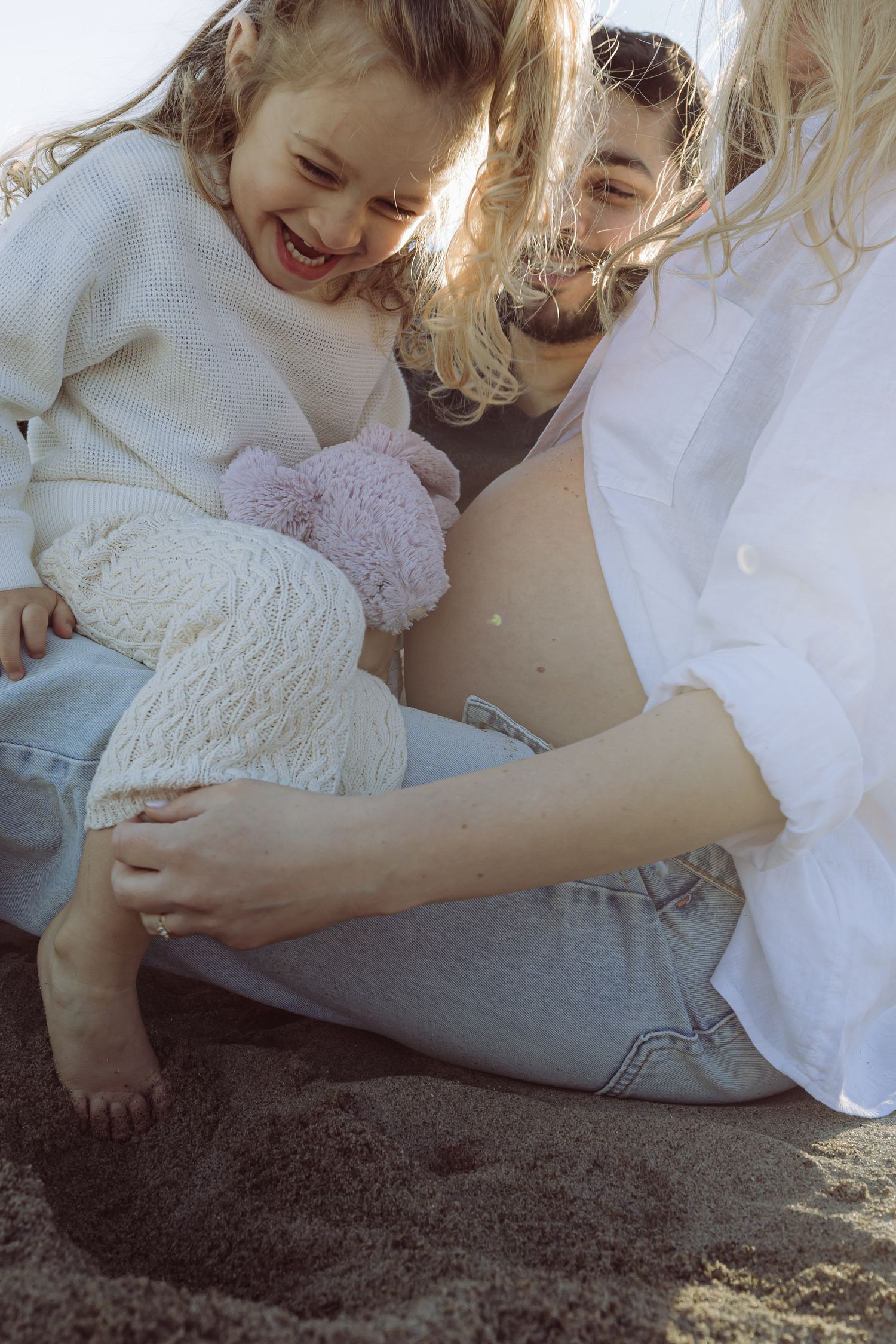A joyful candid moment shared between family members laughing and cuddling with a plush toy in sunlit setting on beach during maternity.