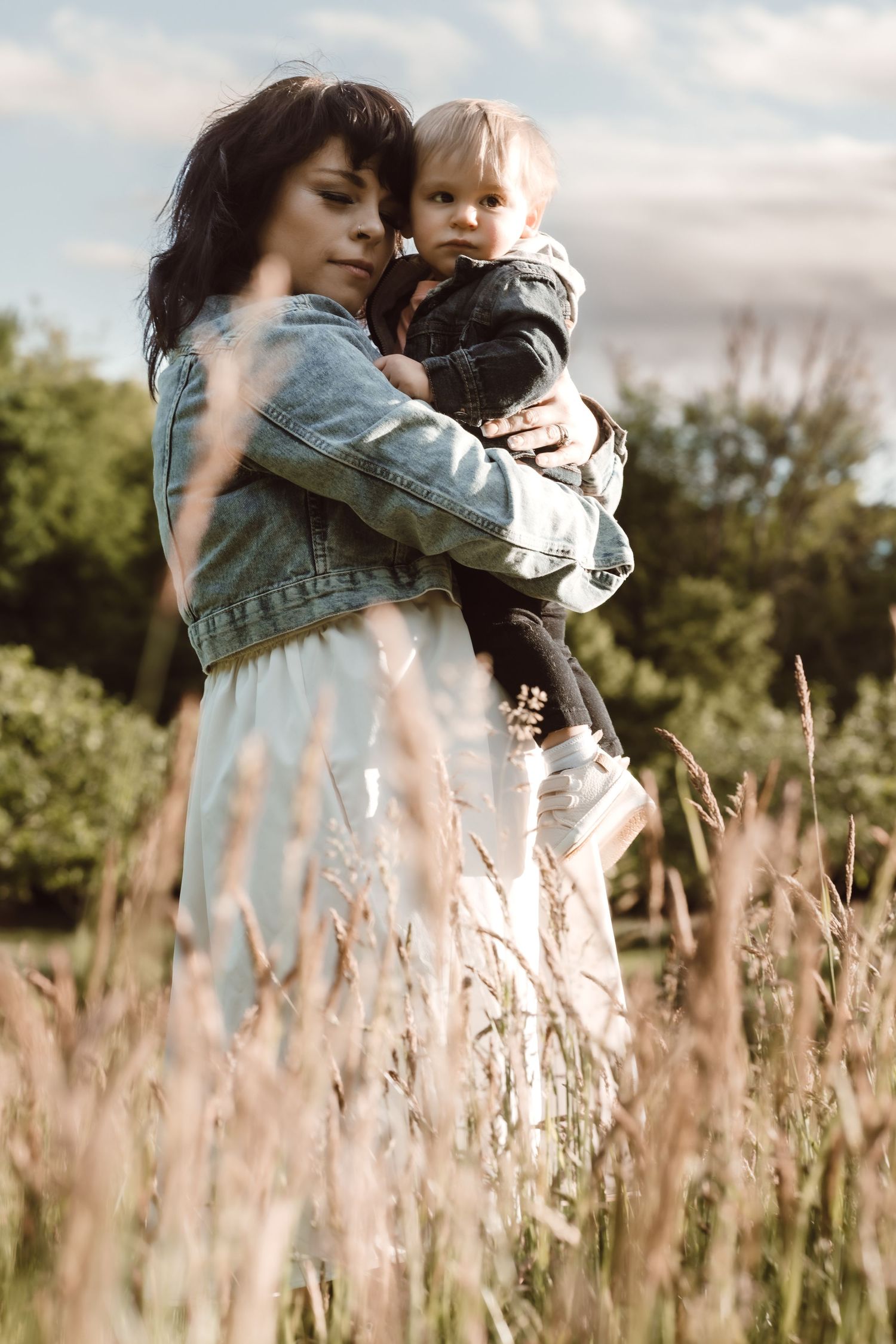 A tender moment captured in a field of tall grass as a parent in denim jacket holds and embraces a small child at sunset.