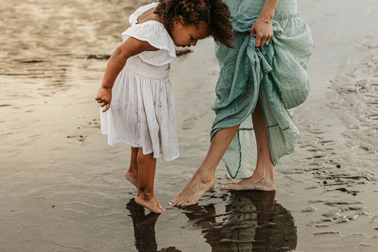 A mother and 2 year old daughter point their feet leaving in the wet sand at sunset. Shadows of their silhouettes in the sand.