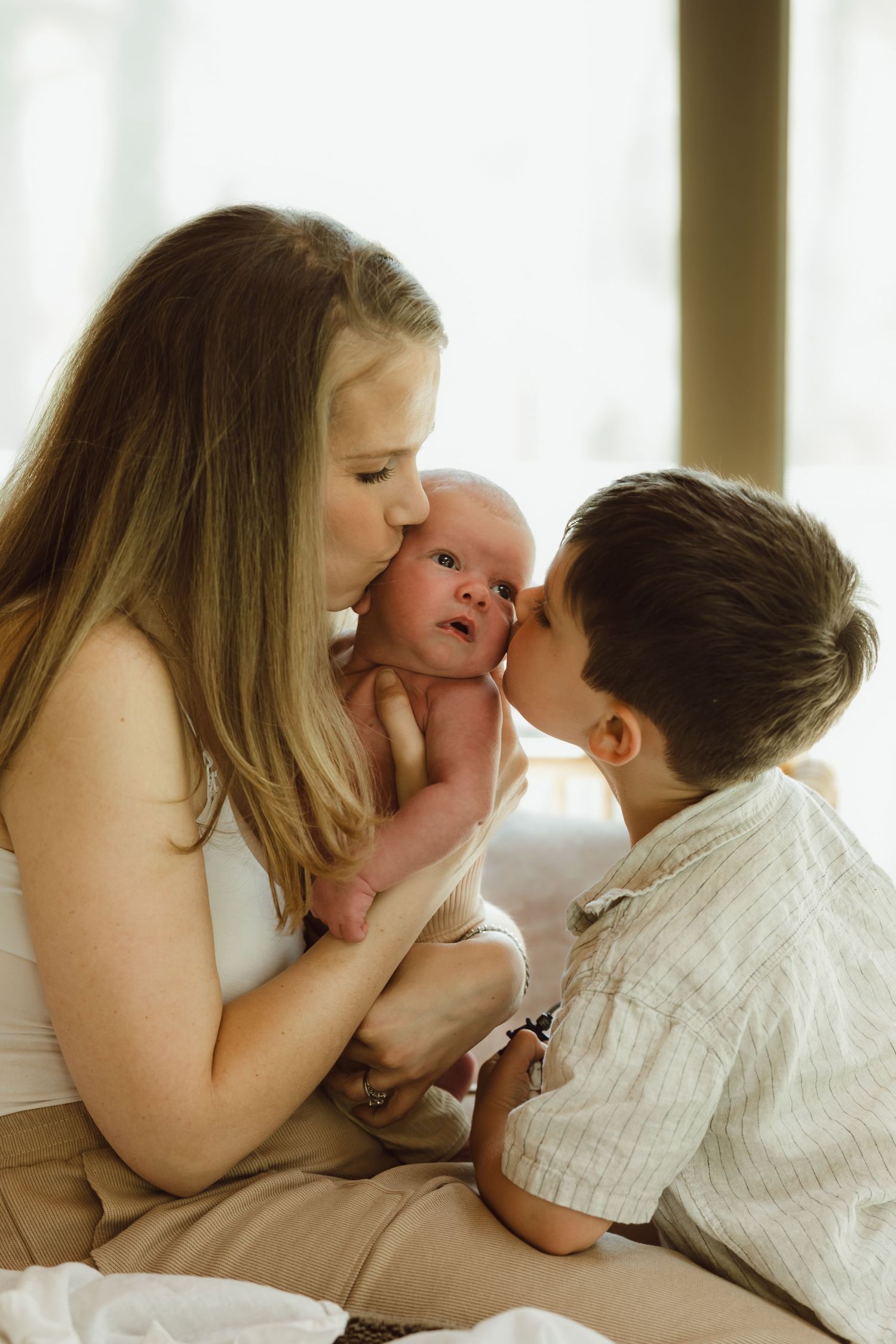 A mother holds her newborn son as she and her toddler son kisses the baby's cheeks for newborn photoshoot in Connecticut.