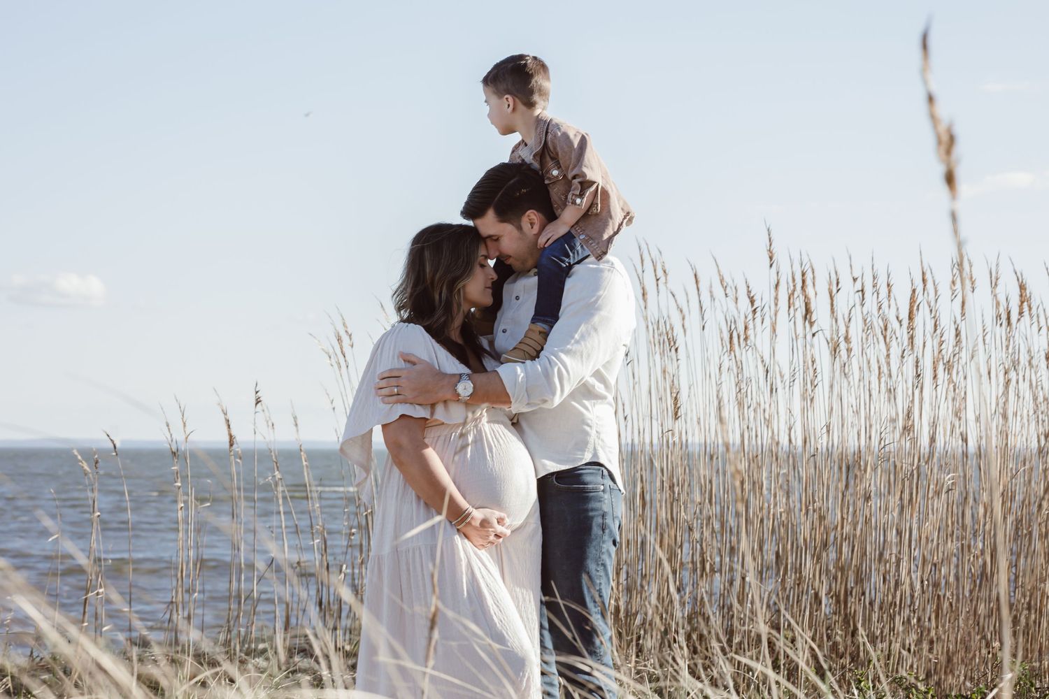 A family embraces tenderly by the ocean with tall beach grass swaying in the breeze. Mother is pregnant and toddler boy on dads shoulders.