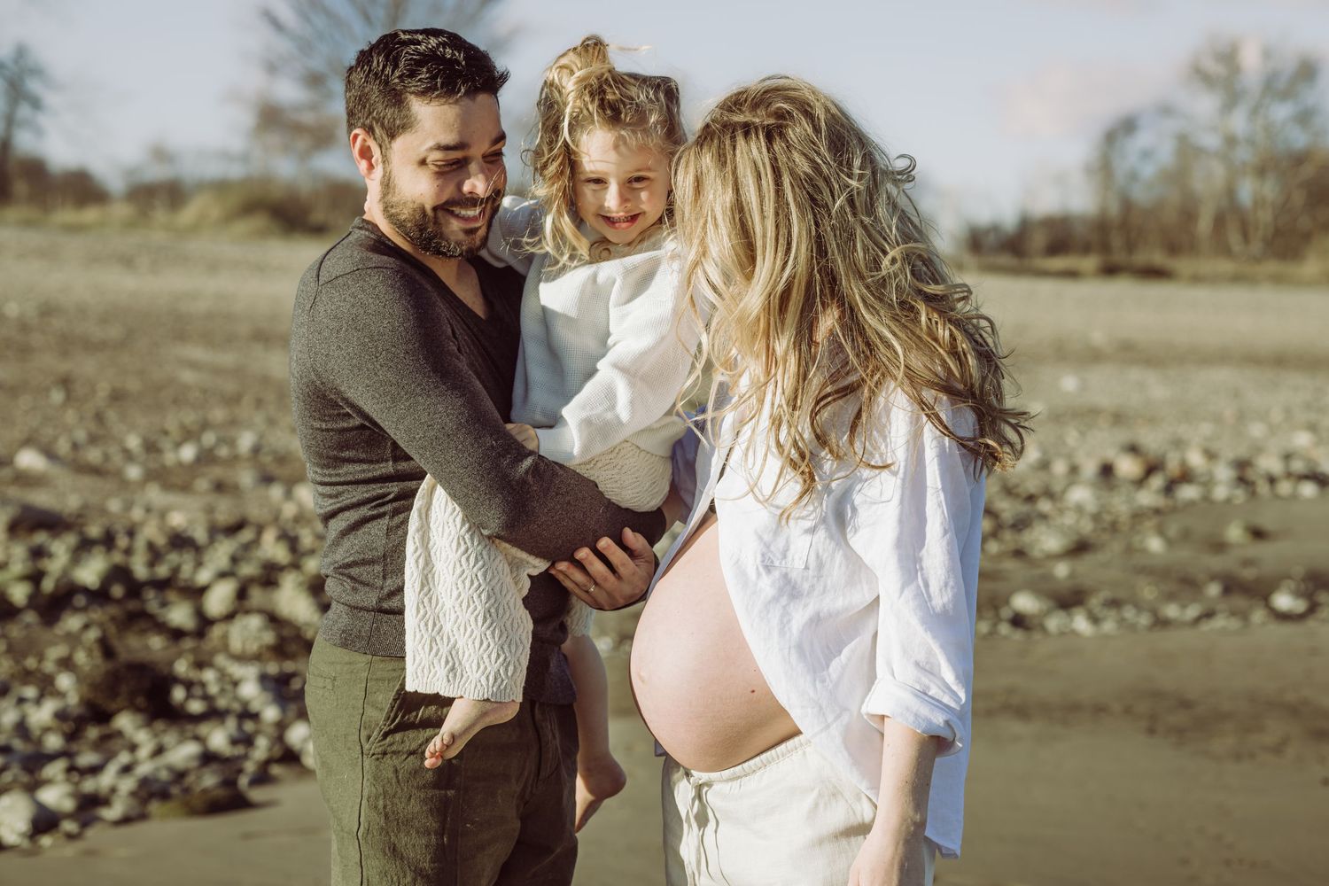 A couple shares a joyful moment with their daughter on a beach while celebrating an expected pregnancy.