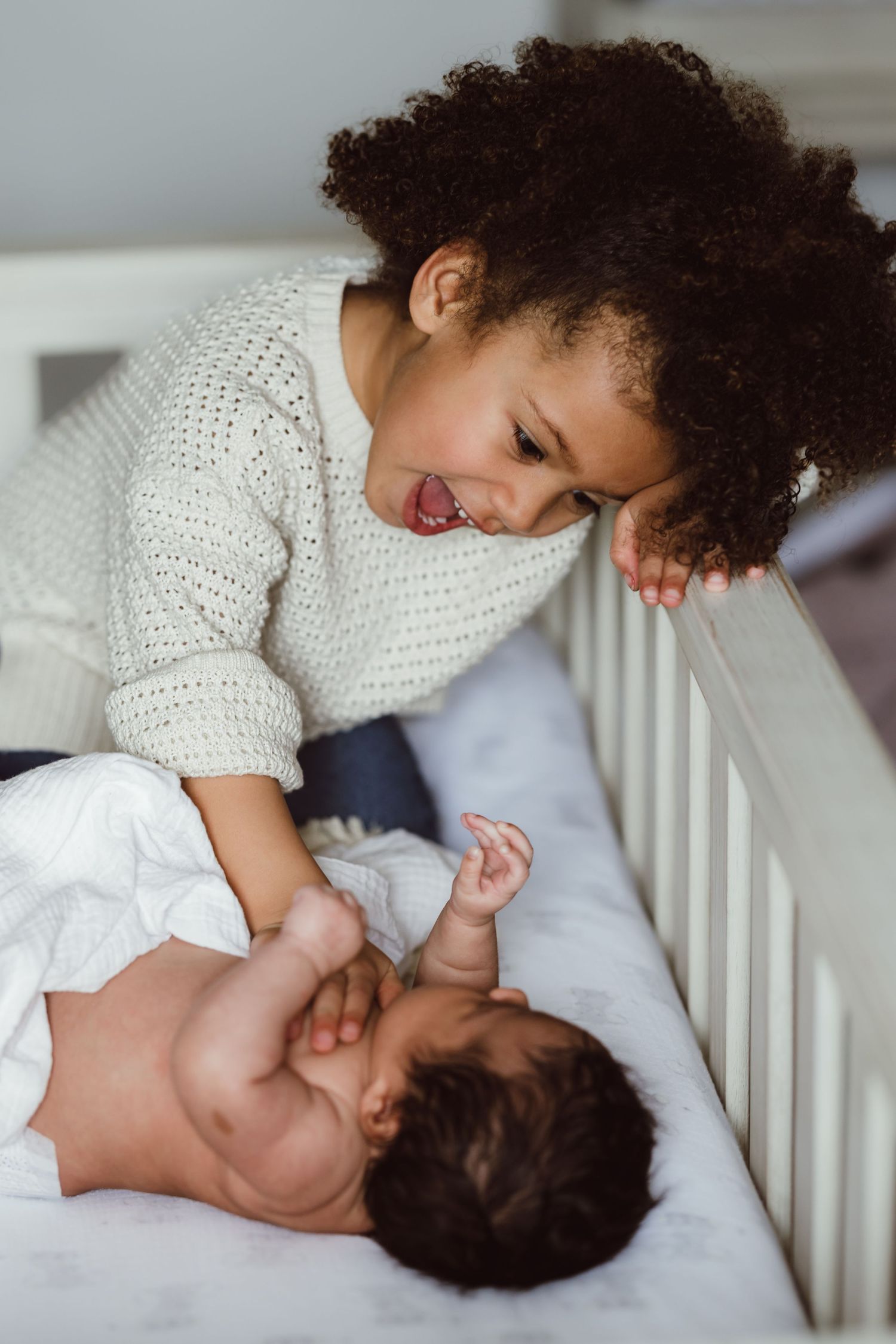 A toddler sister and baby sister in a white crib, showing loving interaction and bonding during newborn photoshoot.