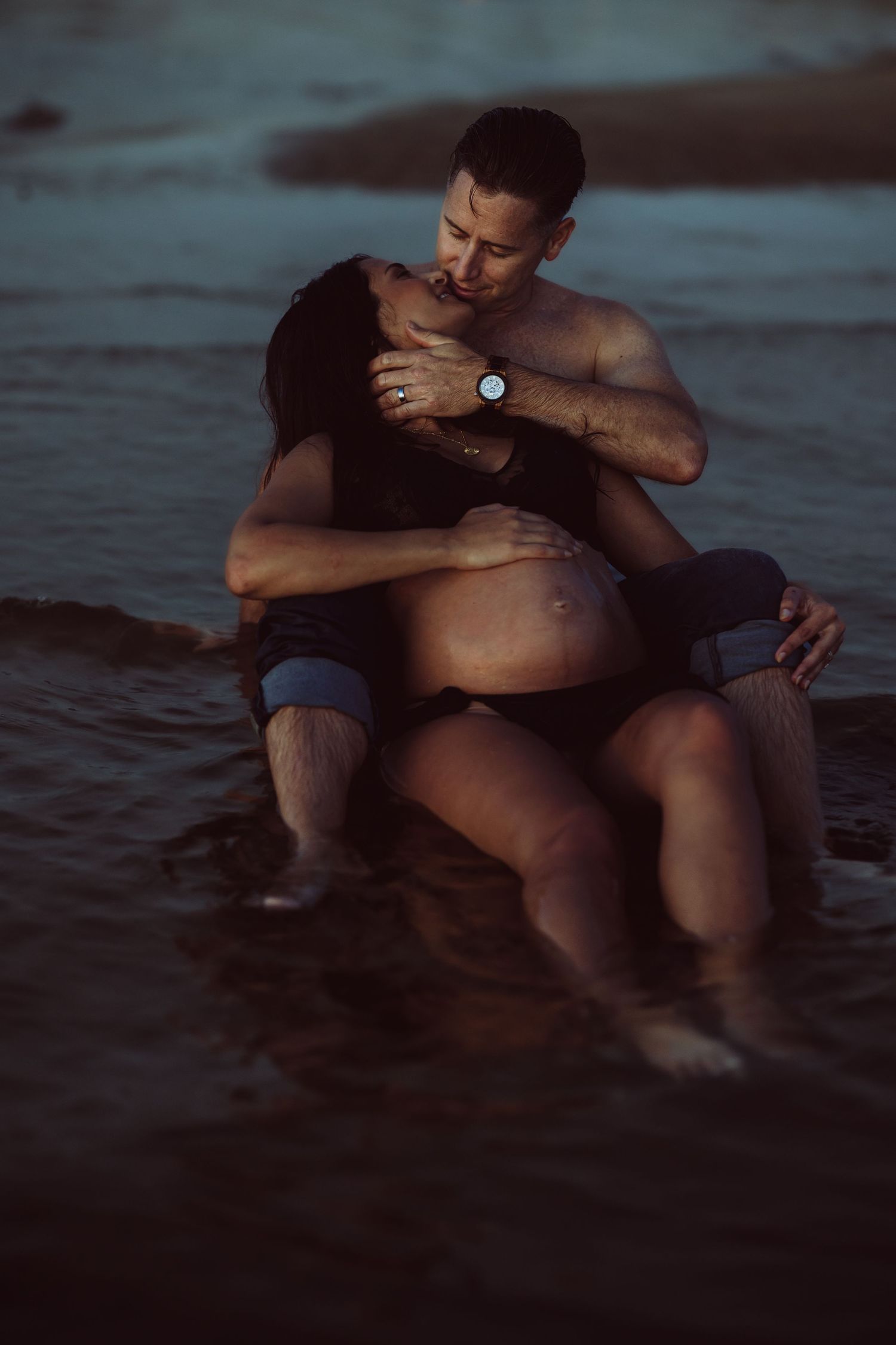 Pregnant woman laying on husband in an intimate moment while sitting in shallow water at dusk for maternity session.