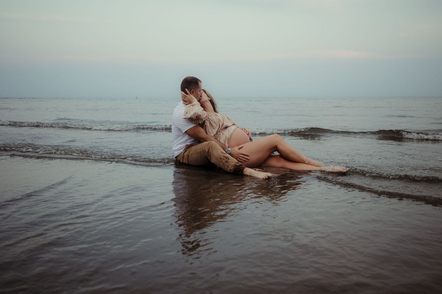 Pregnant woman laying on her husband in the low tide on a beach at sunset with waves gently rolling in around her.