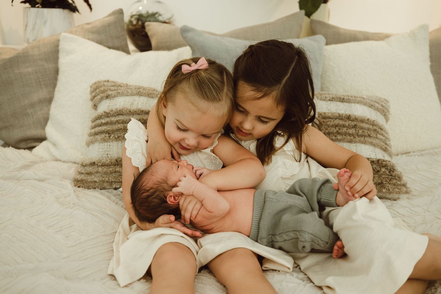 Two toddler big sisters hold their newborn baby brother on a textured white and beige pillowed bed for newborn photoshoot.