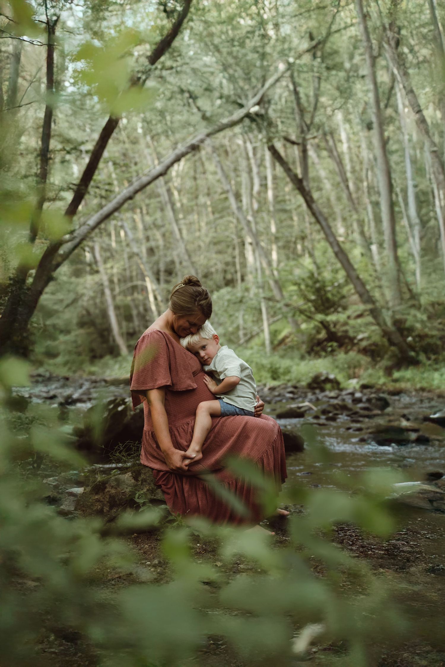 A pregnant mother embraces toddler son while resting together by a peaceful forest stream on a summer afternoon for maternity photoshoot.