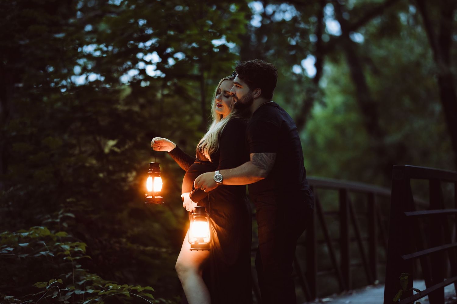 Pregnant woman in wilton connecticut holding glowing lanterns stands on wooden bridge at dusk in moody forest setting.