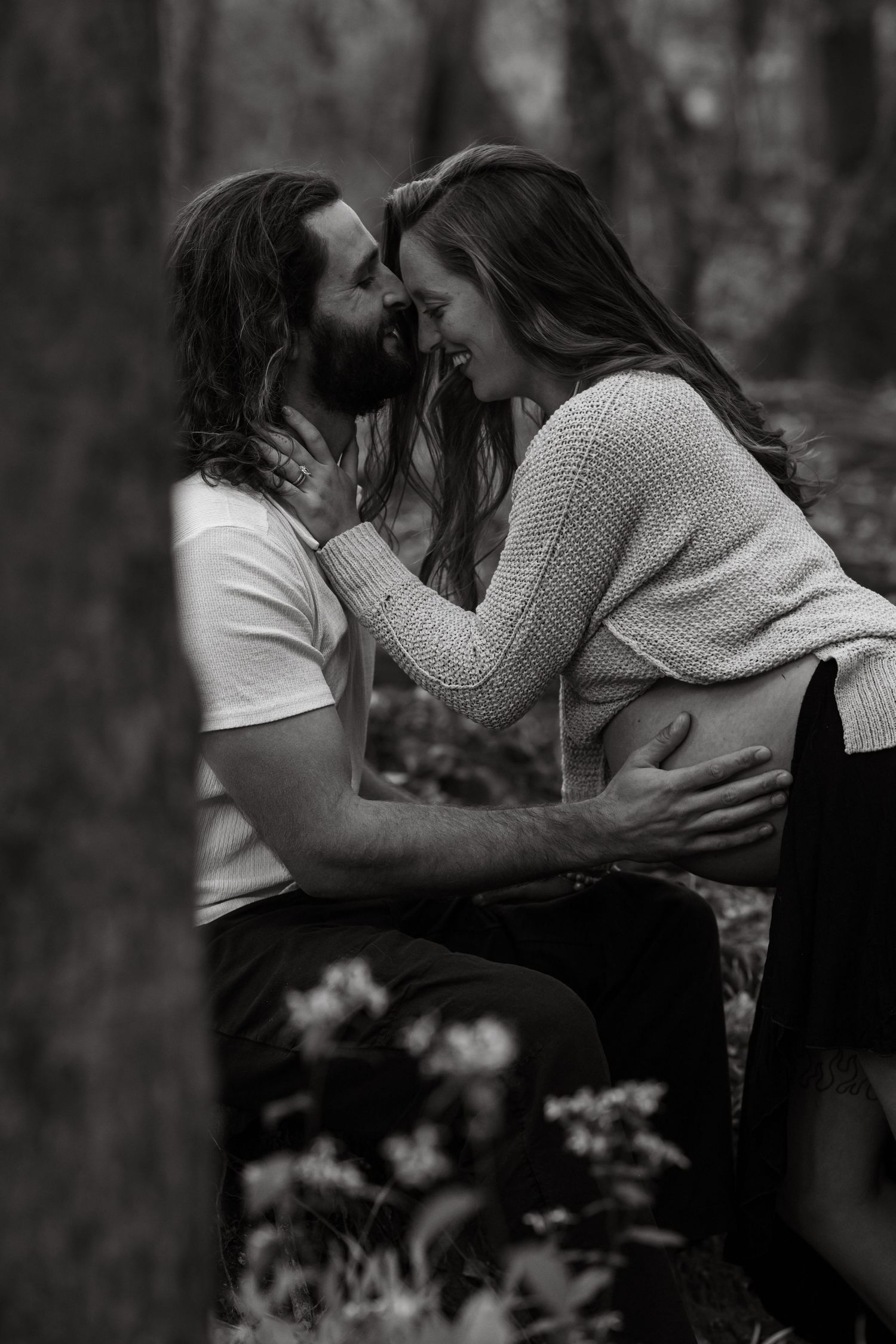A romantic black and white portrait of a couple sharing a moment in a forest setting during a maternity session. Husband hand on belly.