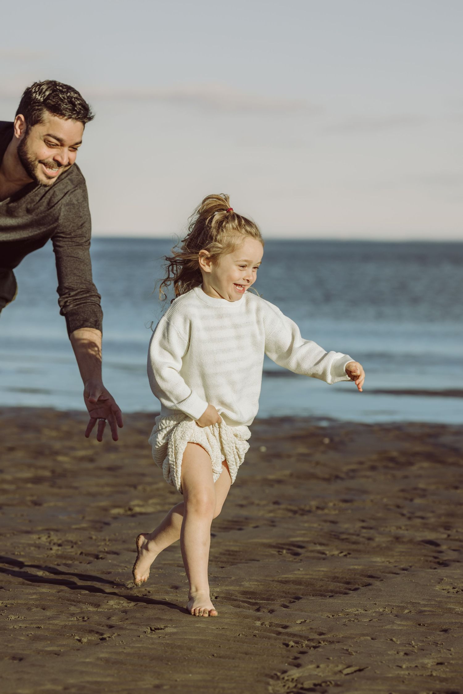 Father and young girl playing together and running barefoot on a sandy beach by the ocean at sunset.