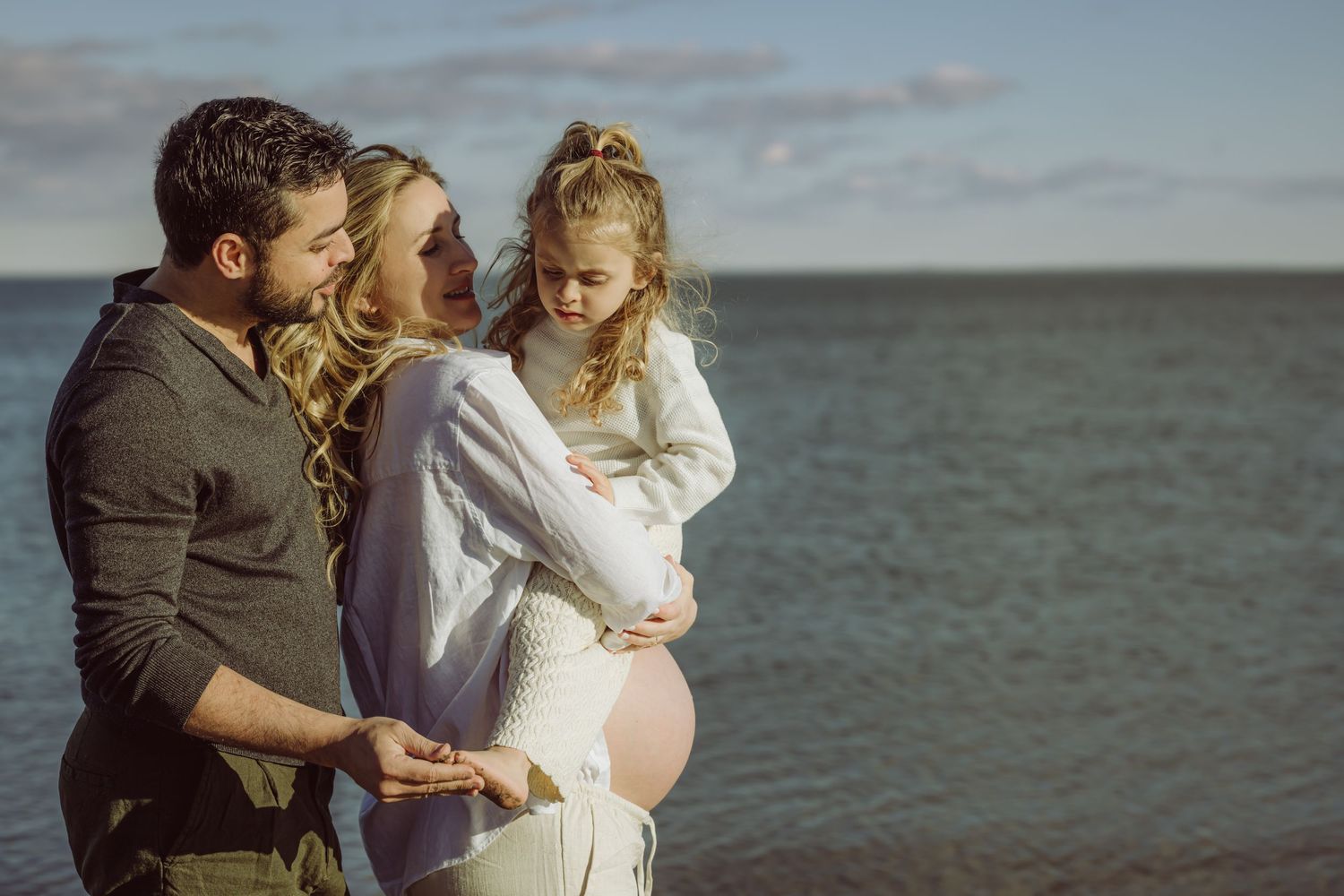 A couple stands at the beach embracing a pregnant woman in a white linen button down and pants by the ocean shore before sunset.