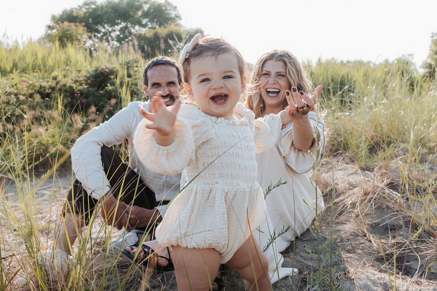 A joyful family wearing white outfits shares laughter while sitting together in a grassy outdoor setting during golden hour in connecticut.