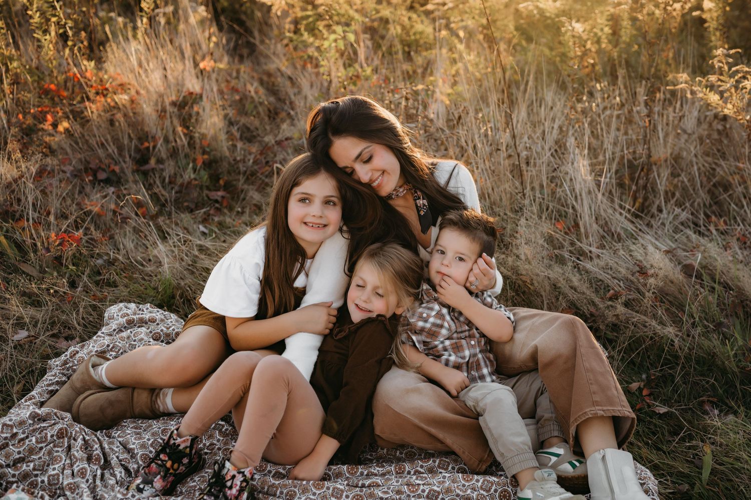 A mother with her three kids sits together on a blanket in a field of golden grass during sunset.
