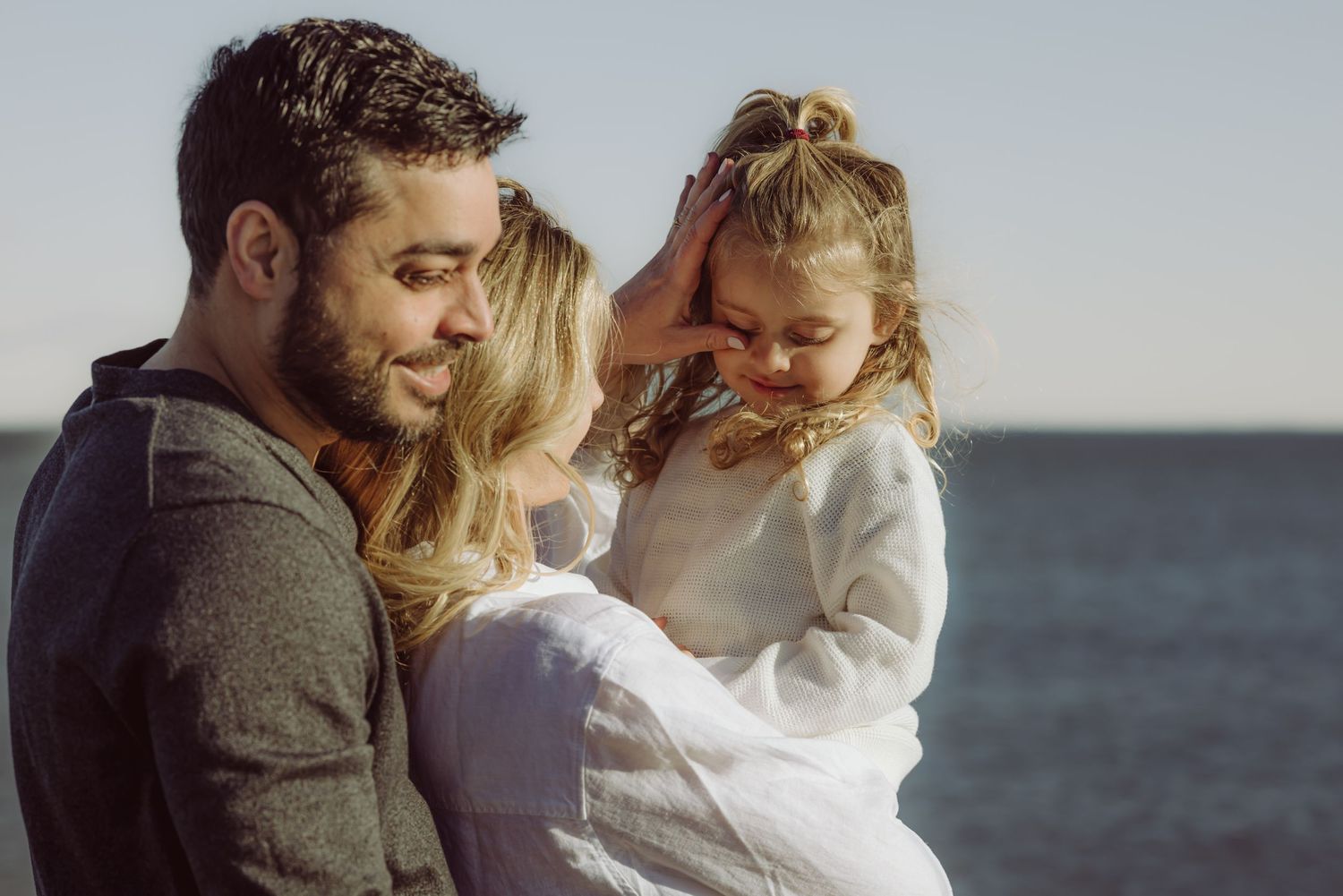 Family enjoys intimate moment together while standing at oceanfront on sunny day with water visible in background.