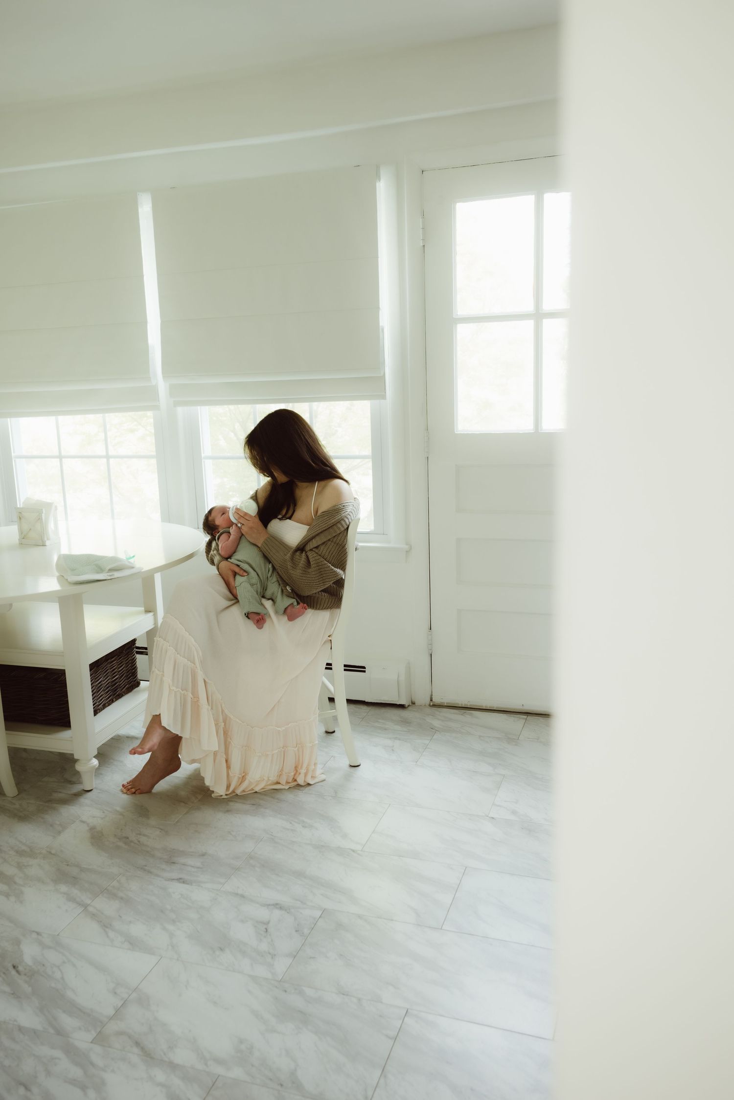 In a bright white kitchen with natural light streaming through windows and white marble flooring a mother bottle feeds newborn son.
