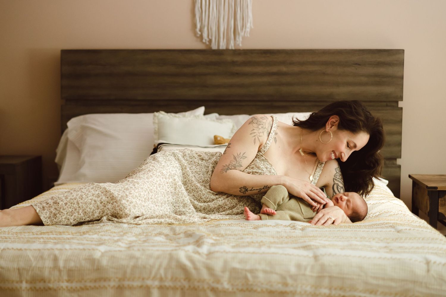 A new CT mom lays in a bed with neutral tones and a macrame wall hanging in the background holding her newborn son for newborn photos.