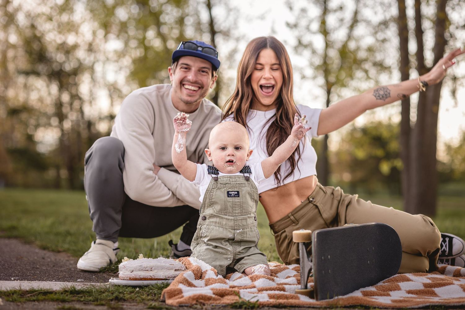 Young parents and one year old son celebrate with hands raised in the air for milestone birthday photoshoot. Cake on boys face and arm.