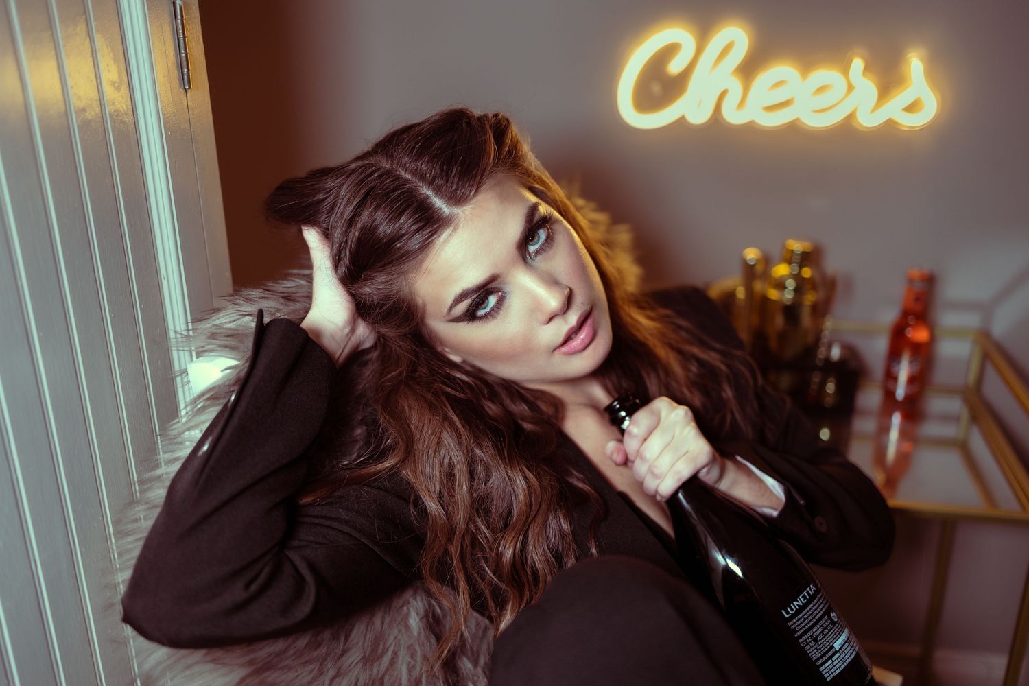 A woman holding a champagne bottle intensly looking at the camera with a sign cheers in the background. Head resting on her hand.