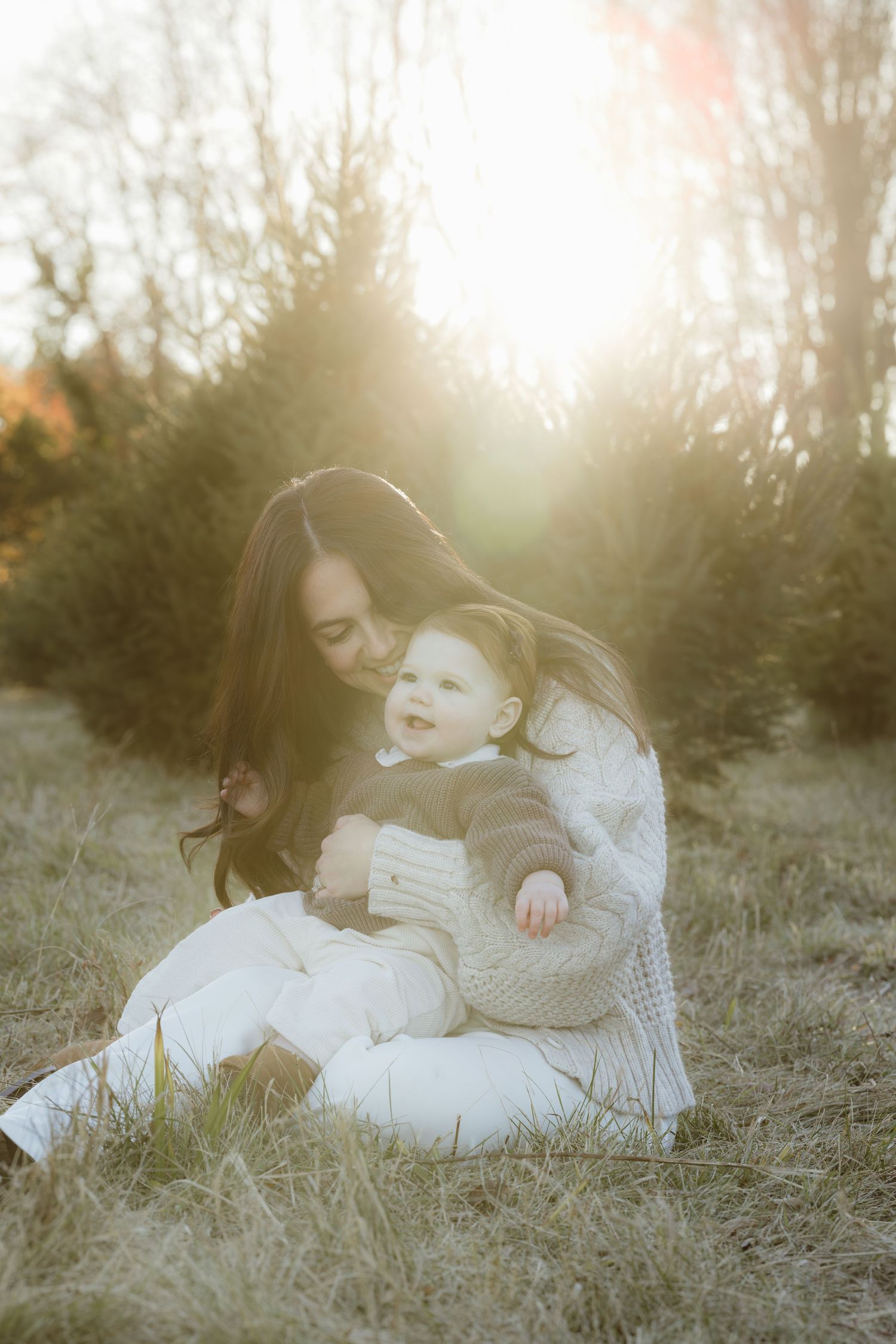 Soft sunlight shines on a mother and her daughter at a tree farm as they snuggle and laugh on the grass.
