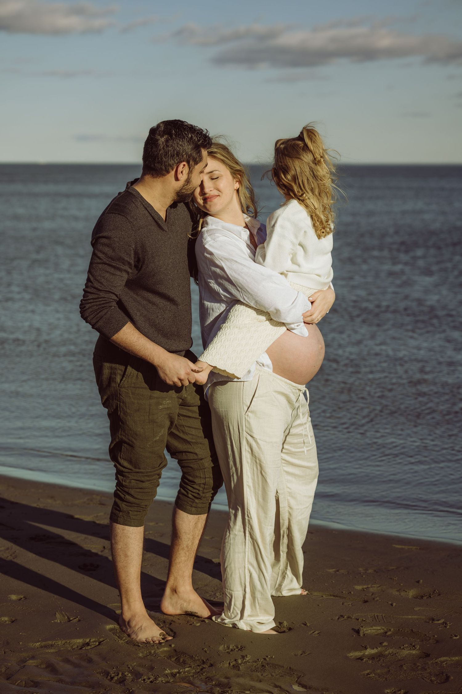 Expectant couple sharing an intimate moment together on a beach at sunset with toddler daughter for maternity photoshoot.