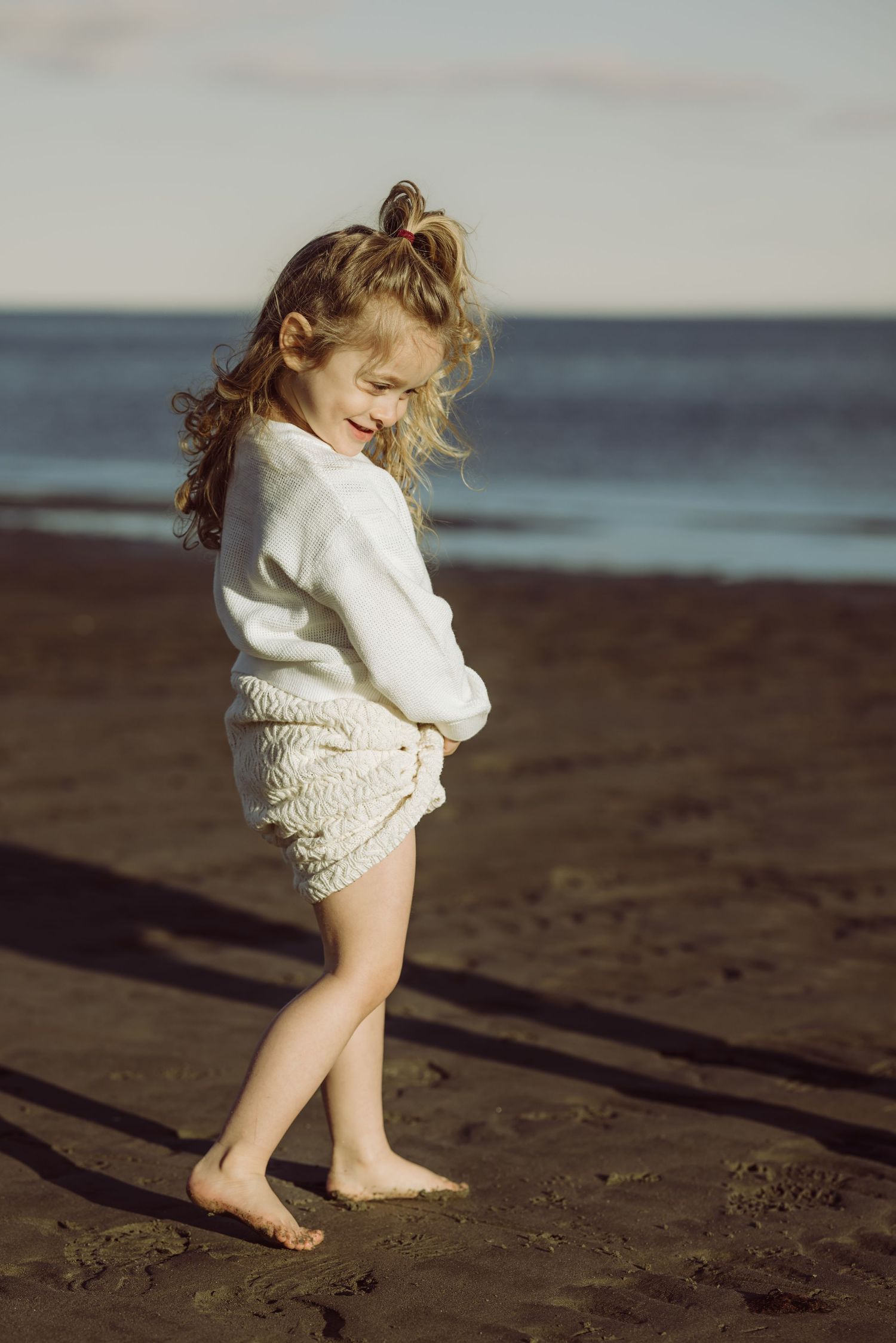 Child in white sweater dress plays joyfully on dark sandy beach at sunset.
